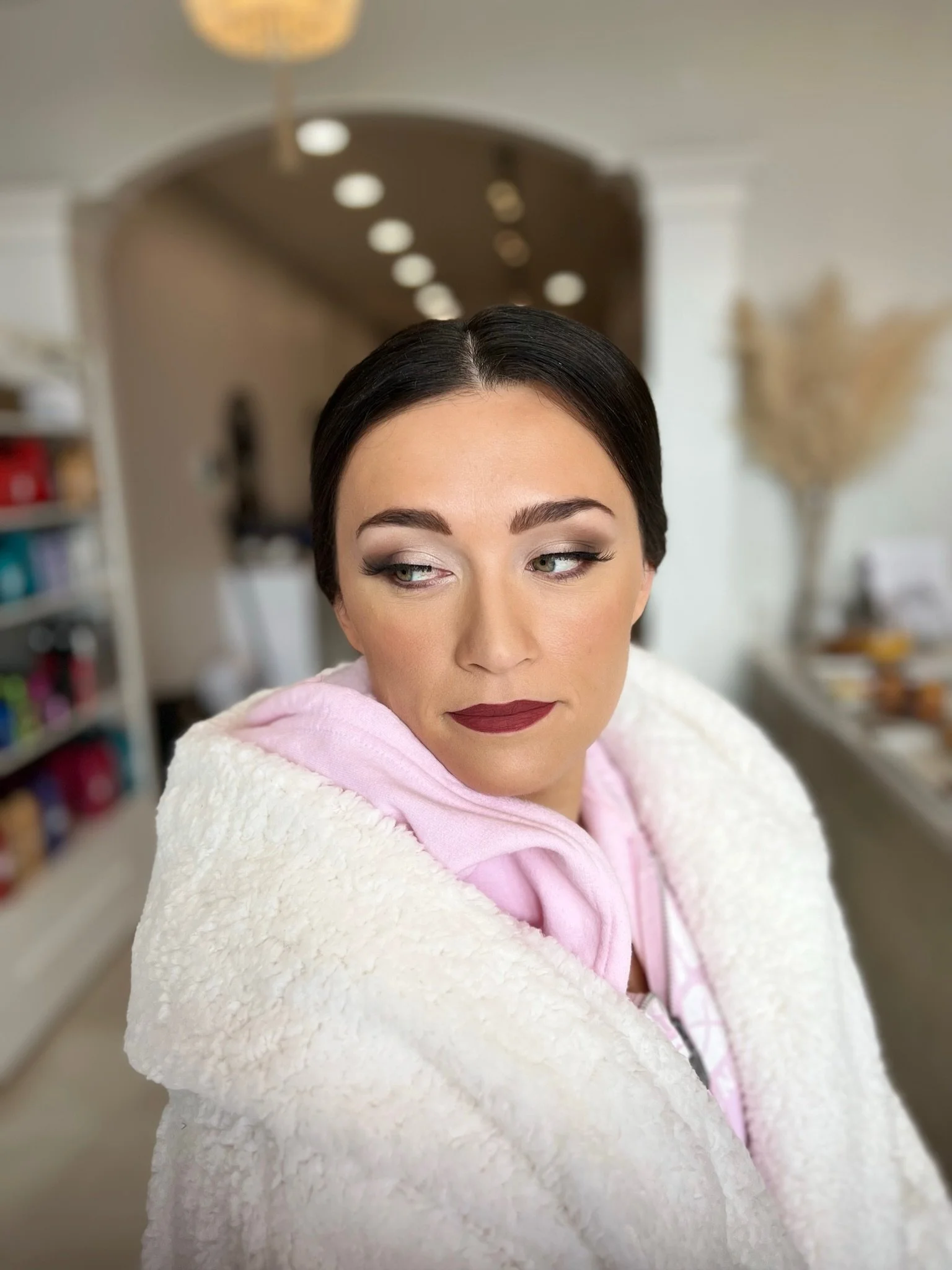 A woman with dark hair styled in a sleek bob, wearing makeup with dark eyeshadow and lipstick, wrapped in a cozy white blanket and a pink hoodie, standing in a home interior with blurred background including shelves and a decorative vase.
