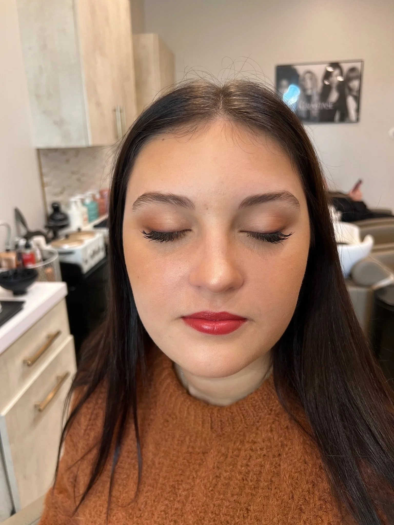 A woman with closed eyes and makeup in a salon or beauty studio. She has long dark hair, well-groomed eyebrows, and red lipstick. The background shows salon equipment and a poster on the wall.