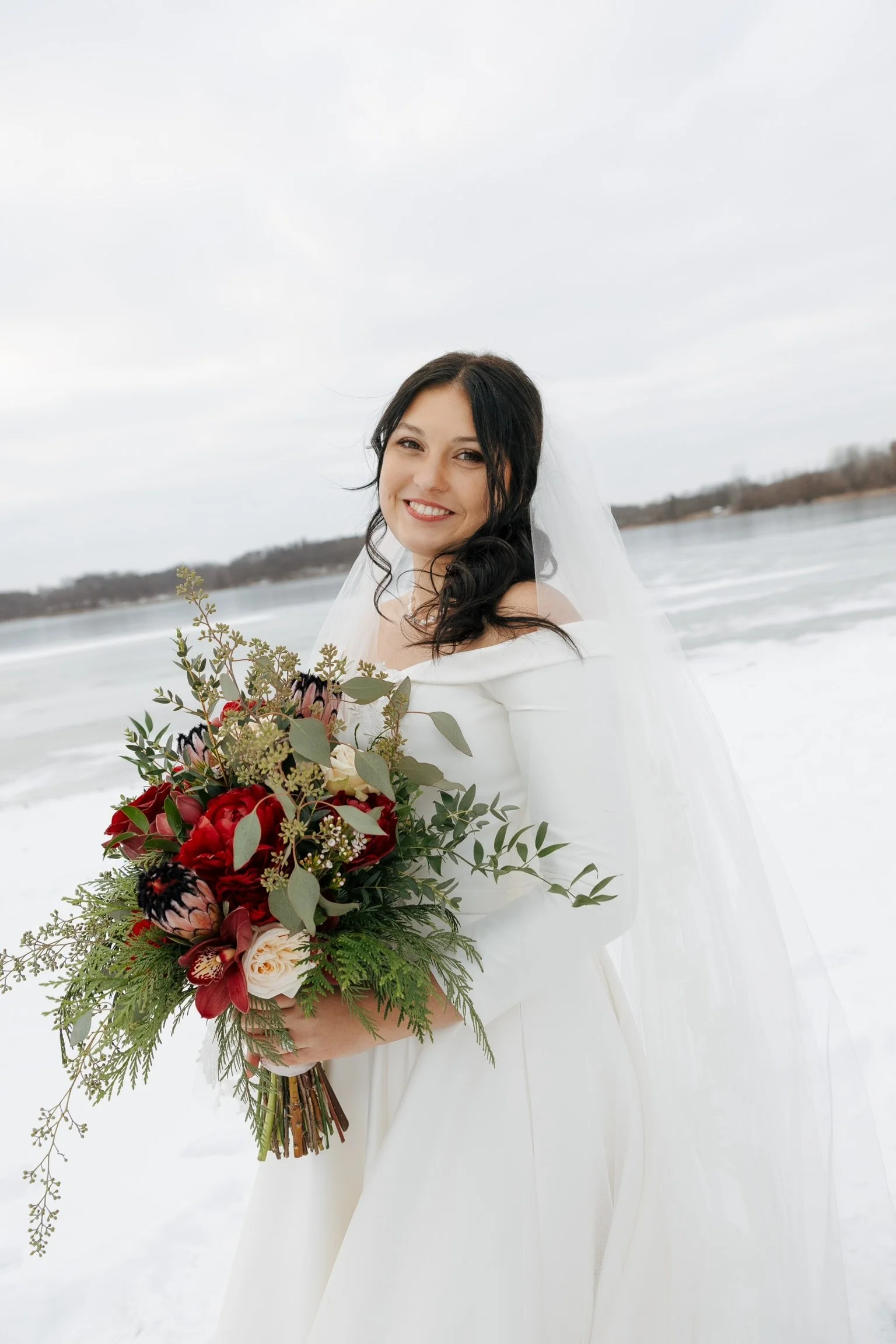 A smiling bride in a white wedding dress holding a large bouquet of red, pink, and white flowers with greenery, standing on a snowy beach with water and trees in the background.