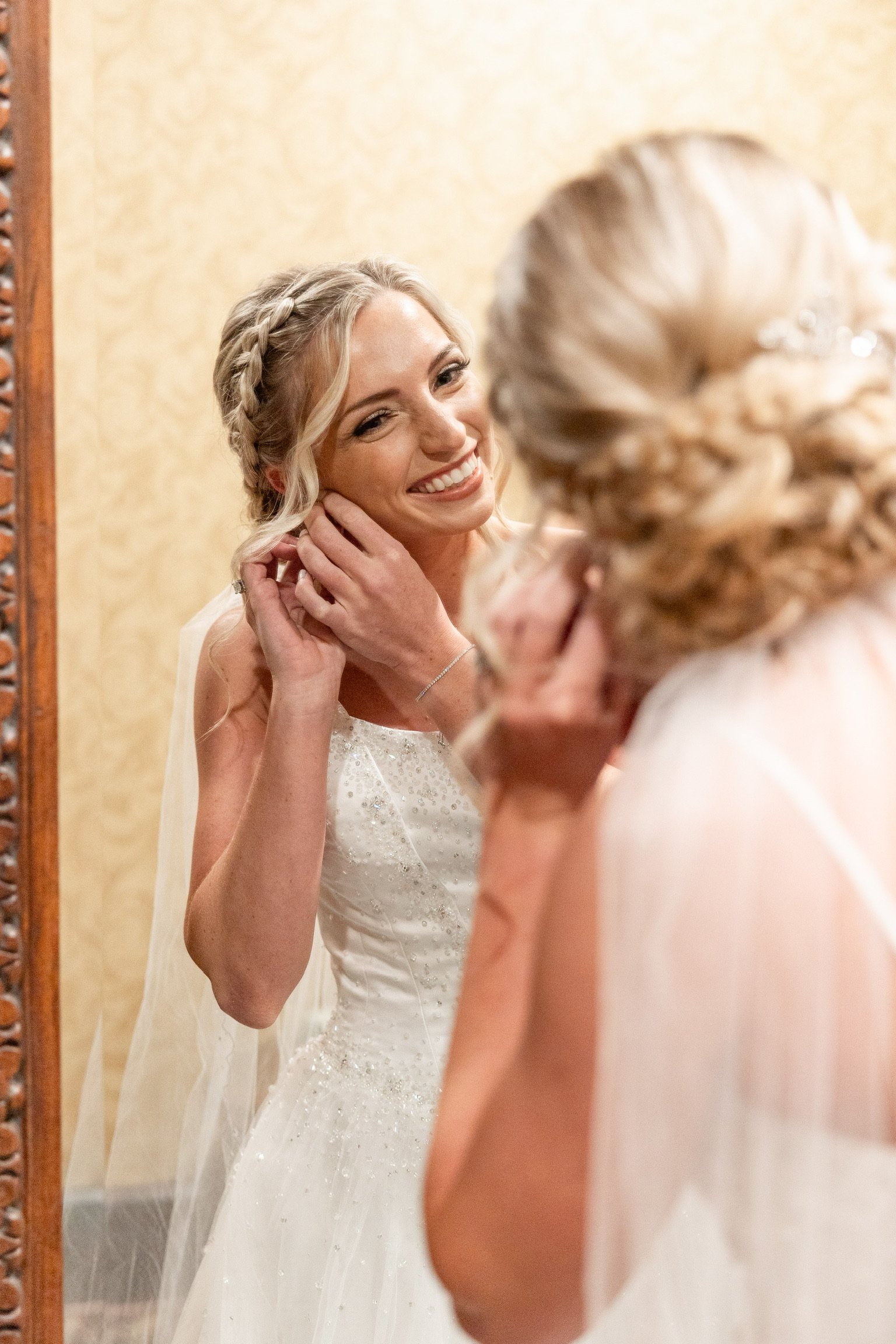 A smiling woman in a wedding dress looking at herself in a mirror, adjusting her earring, with her reflection visible.