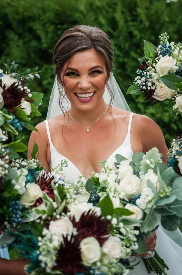 Smiling bride in white wedding dress with veil, surrounded by lush floral arrangements of white roses, blue berries, and greenery outdoors.