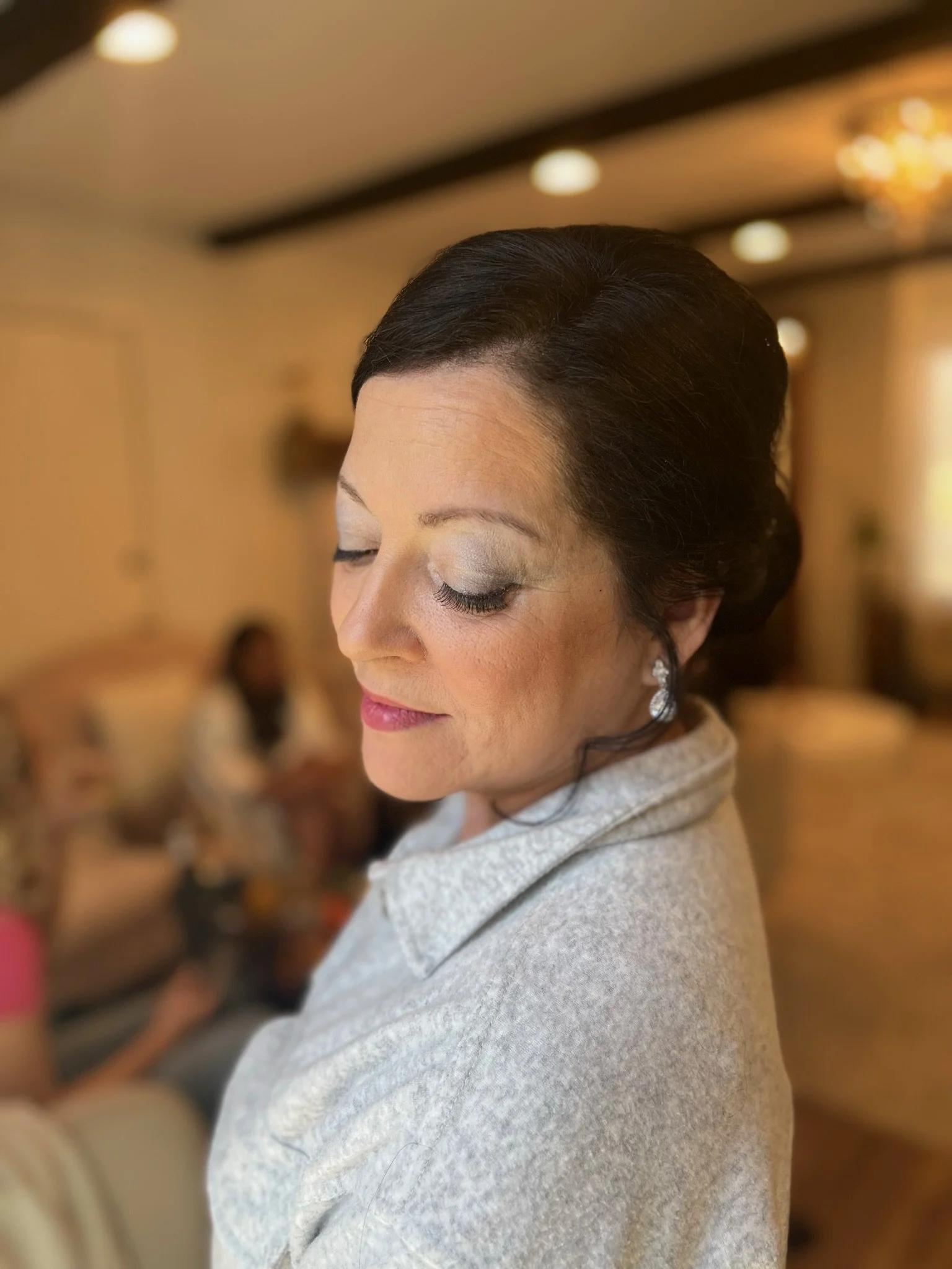 Close-up of a woman with dark hair styled in an elegant updo, with makeup and earrings, in a warmly lit room with blurred background.