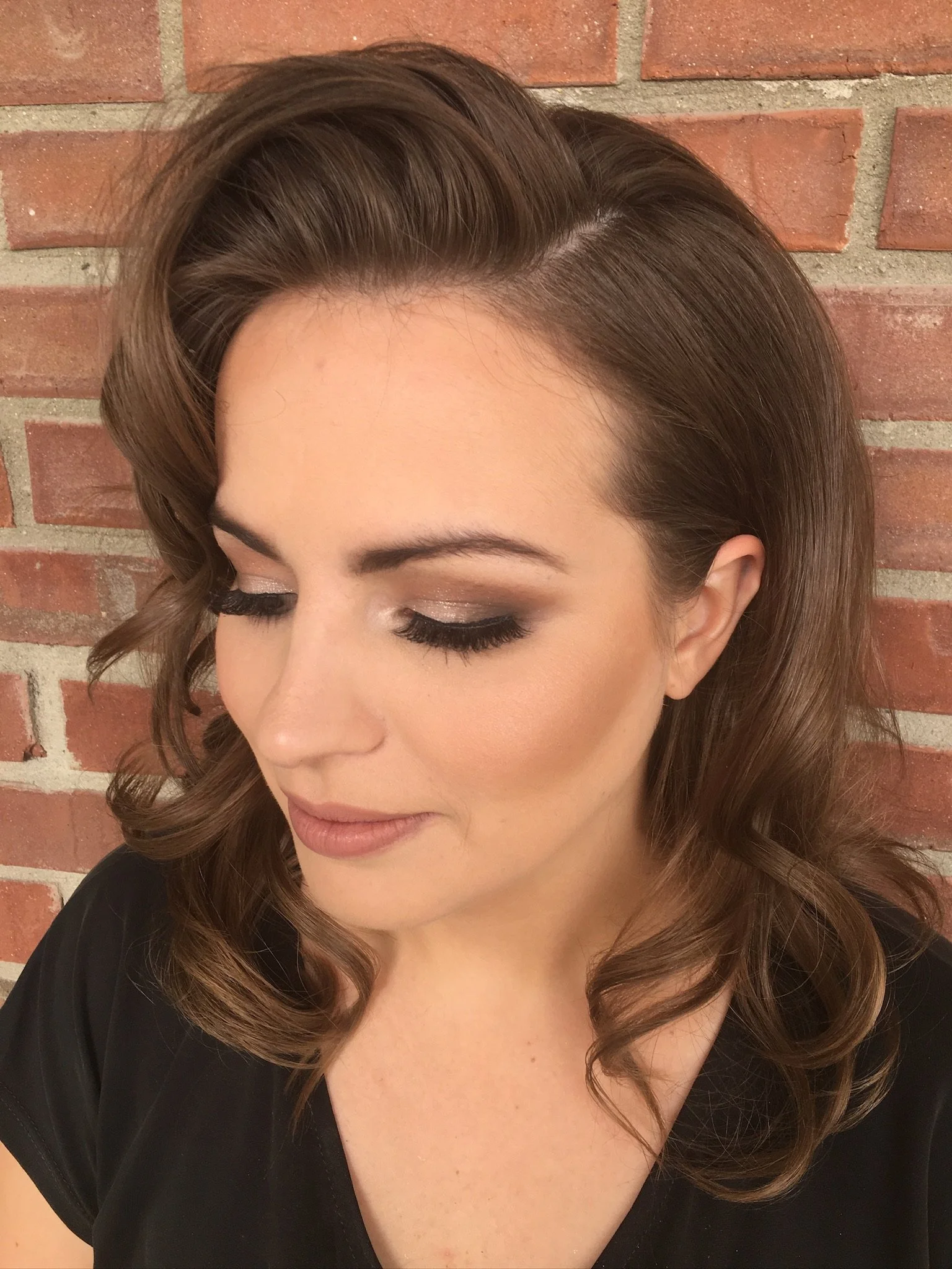 Close-up of a woman with brown hair styled in soft curls, standing against a brick wall background, with her eyes closed and makeup including eyeshadow and mascara.
