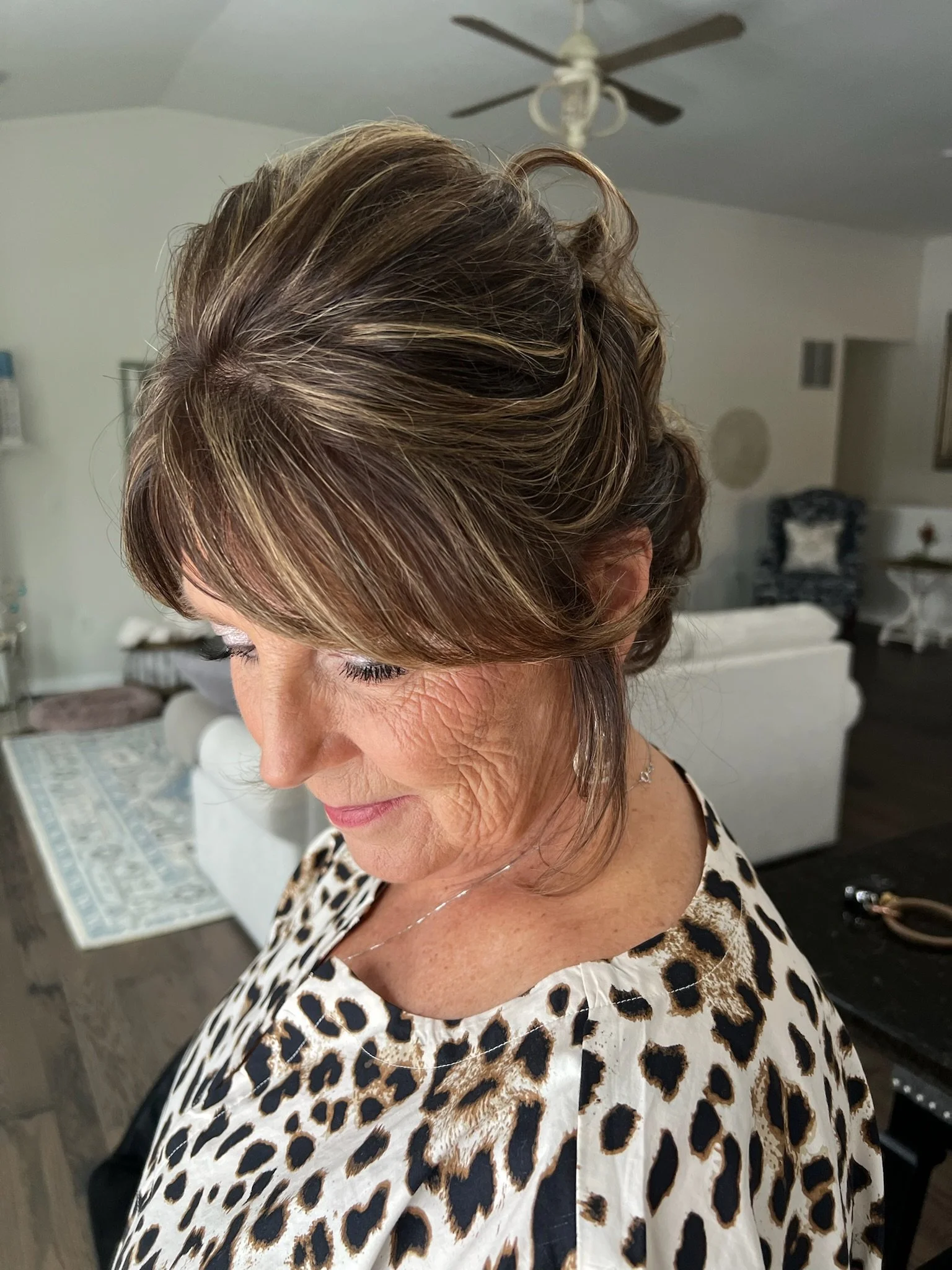 A woman with a short, layered hairstyle featuring highlights, wearing a leopard print top. She is indoors in a room with neutral-colored walls, a ceiling fan, and a chair with a decorative pillow in the background.