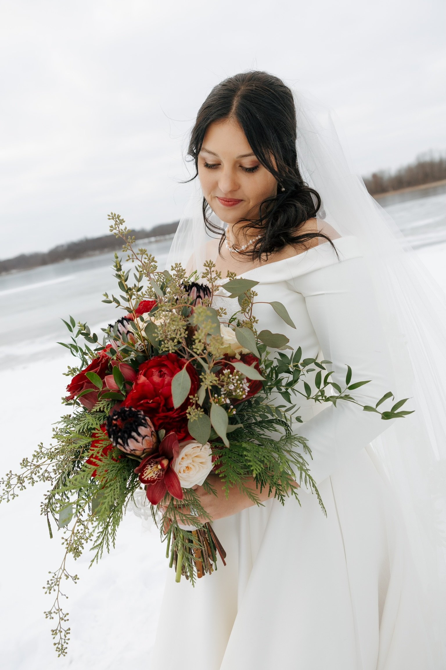 A bride holding a bouquet of red and white flowers with greenery, standing on a snowy landscape with a cloudy sky in the background.
