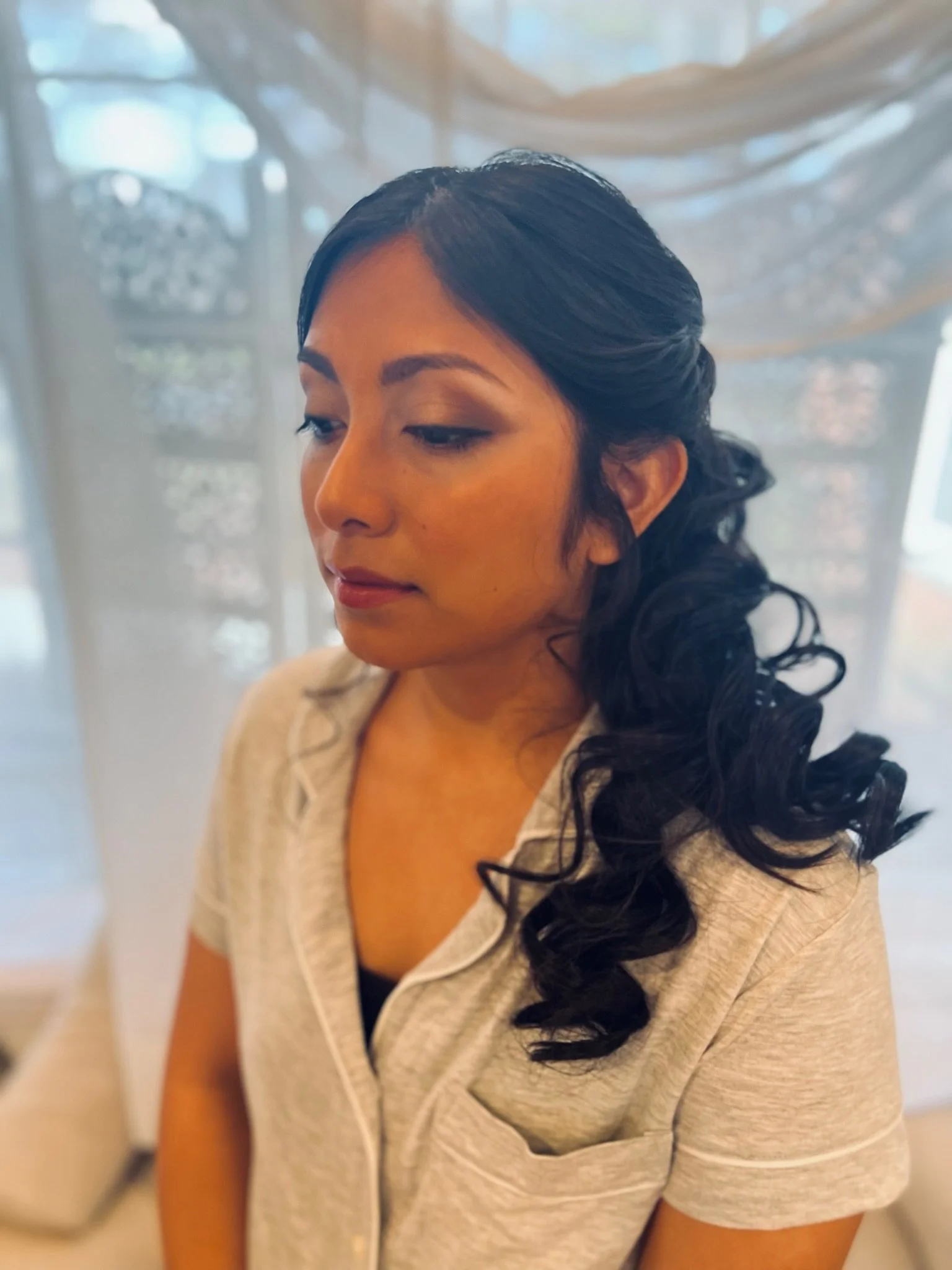 Close-up of a woman with dark curly hair and makeup, wearing a light-colored zip-up sweater over a black top, sitting indoors with a soft-focus background.