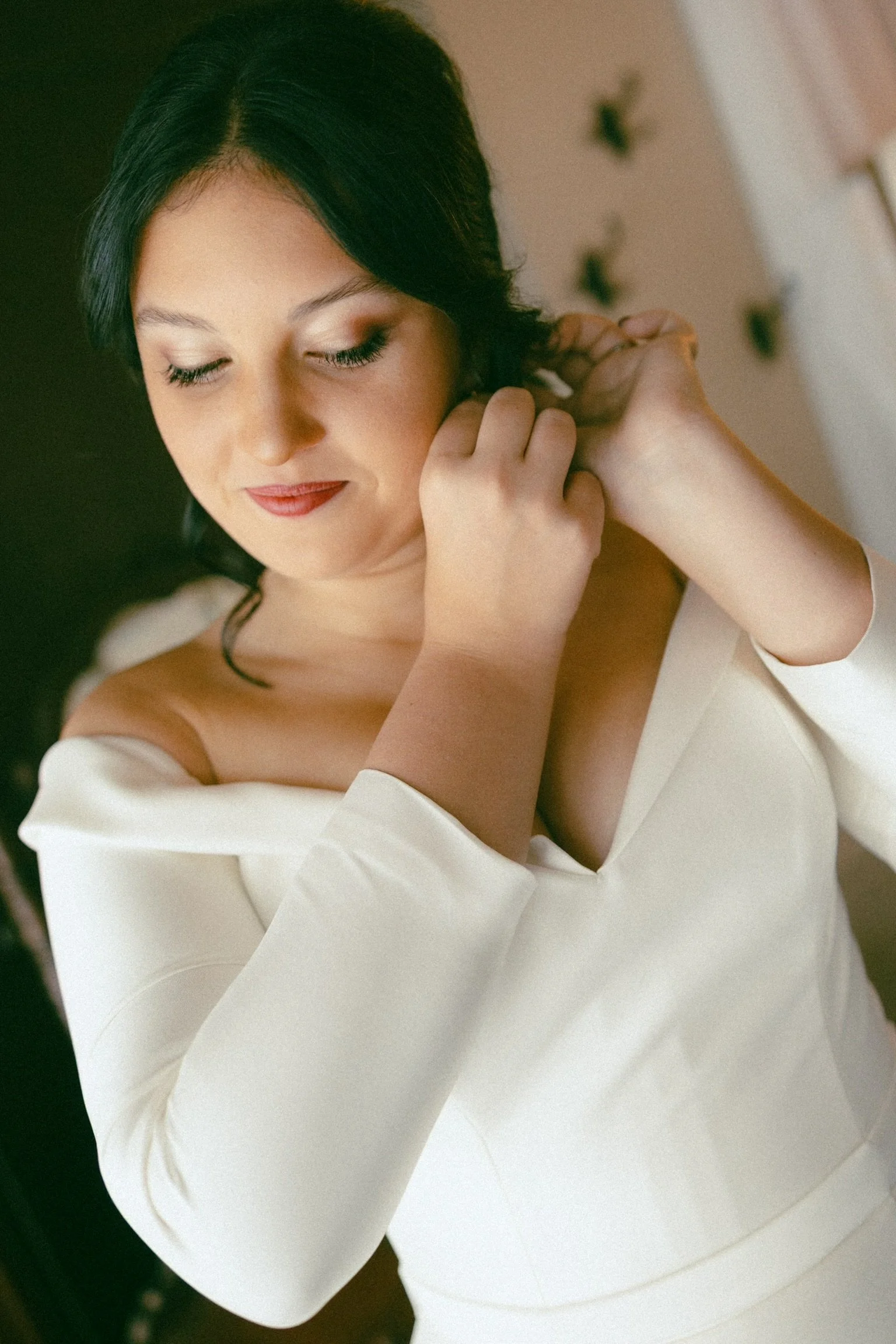 A woman with black hair and makeup putting on earrings, wearing a white off-shoulder top, smiling softly with eyes closed.