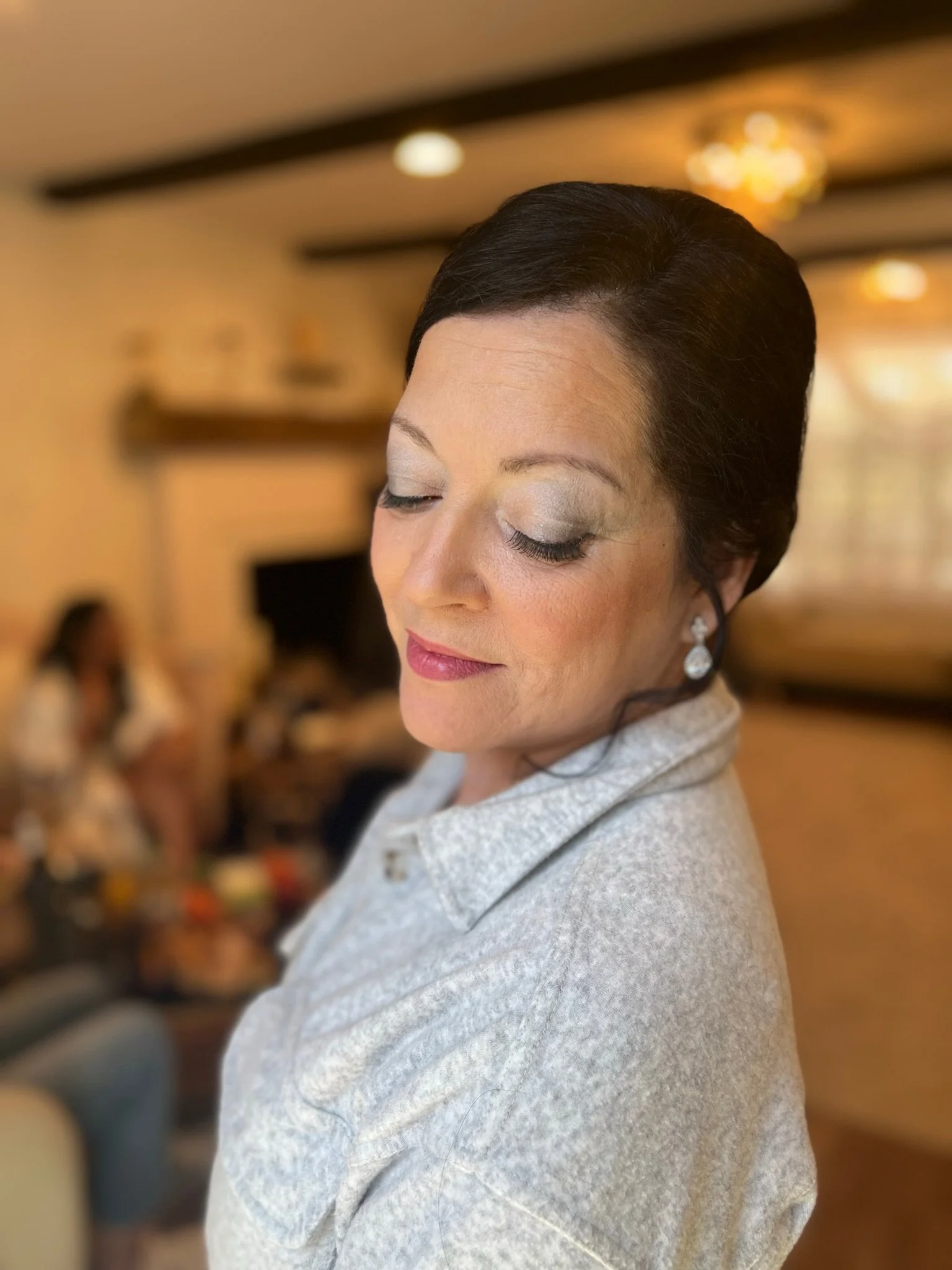 A woman with short dark hair, closed eyes, wearing makeup, earrings, and a light-colored sweater, in a room with blurred background showing people and warm lighting.