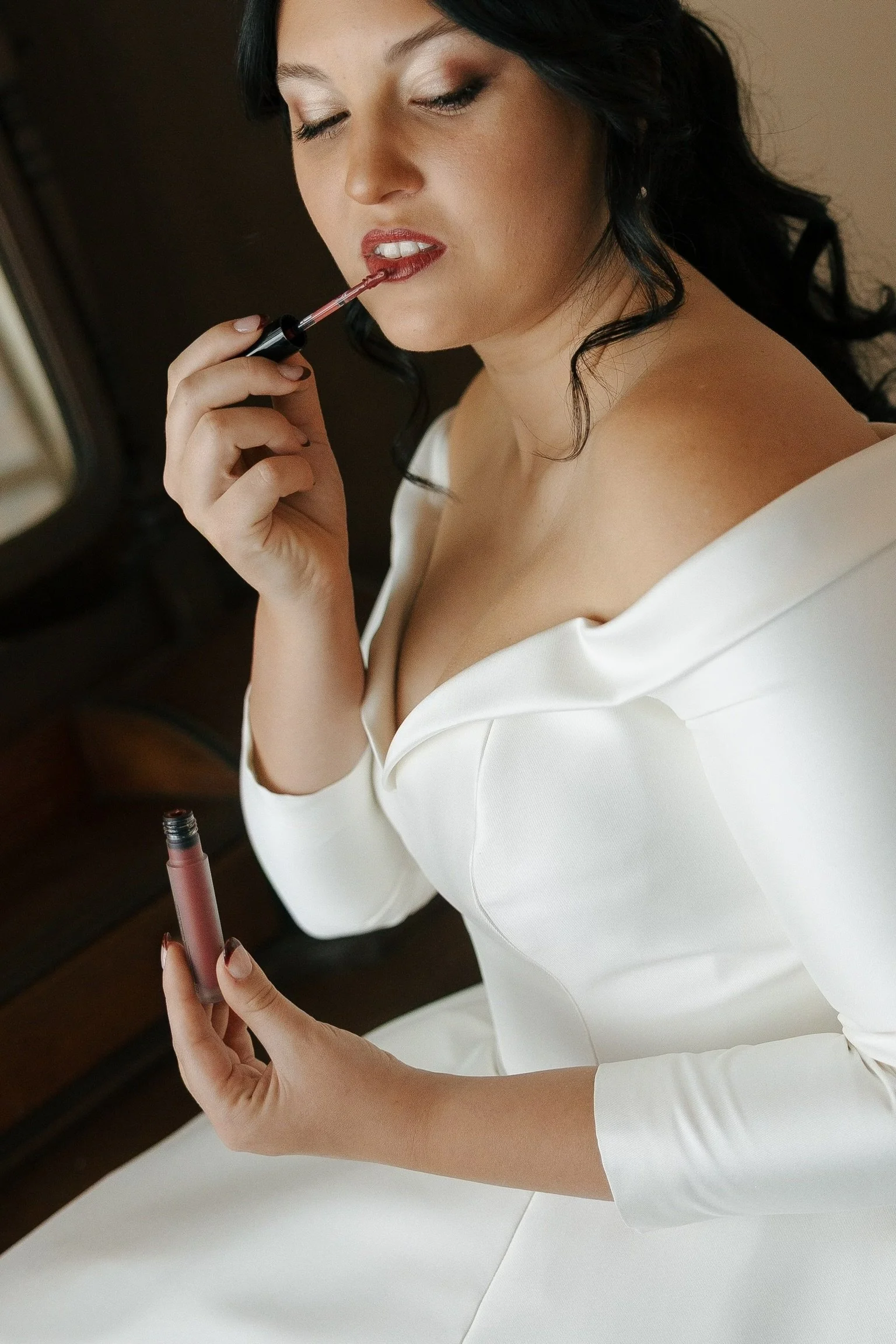 A woman with black hair applying lipstick with a lip gloss wand, wearing a white off-the-shoulder top.