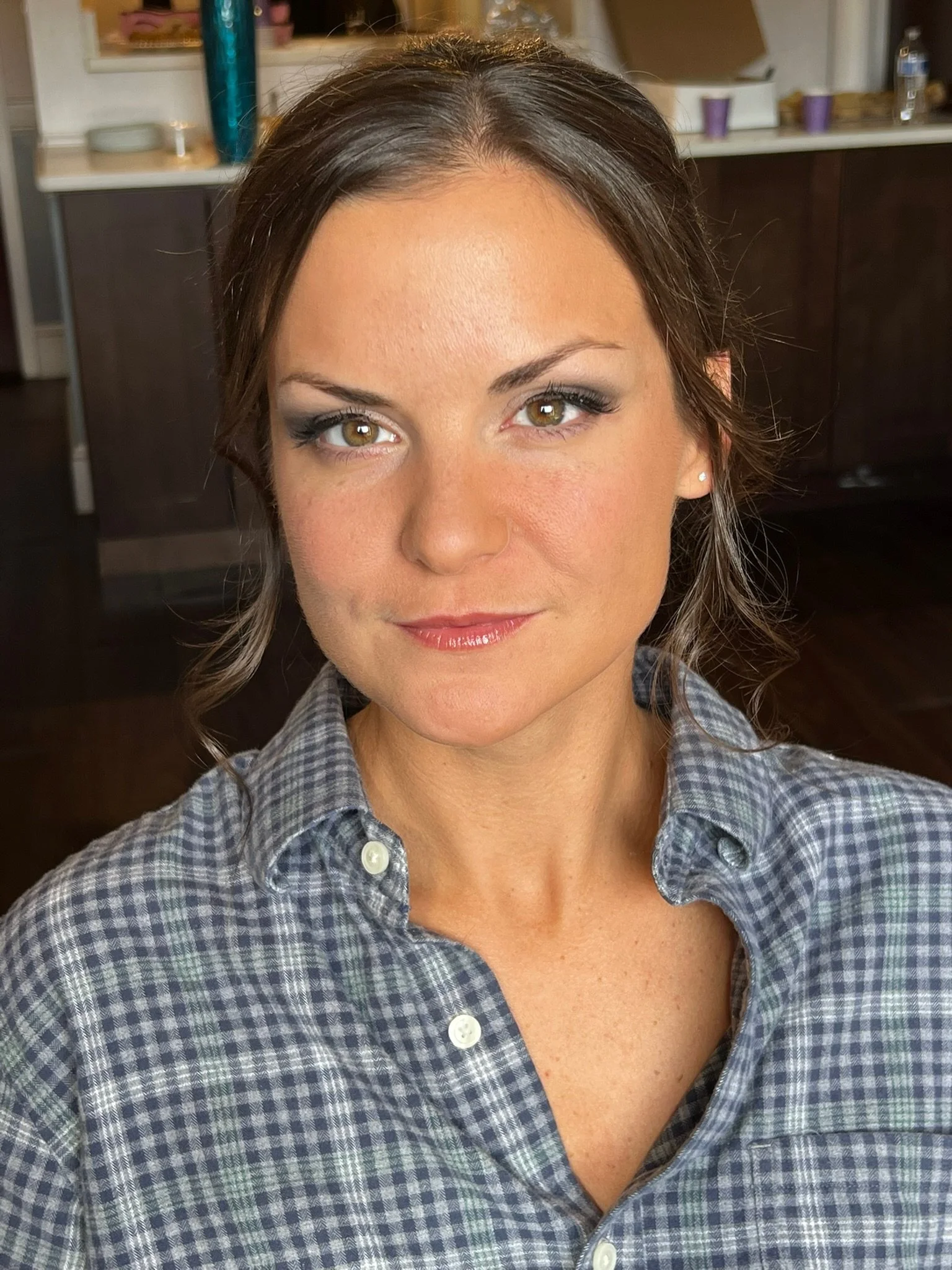 A woman with brown hair and green eyes wearing a plaid button-up shirt in an indoor setting with shelves and kitchen items in the background.