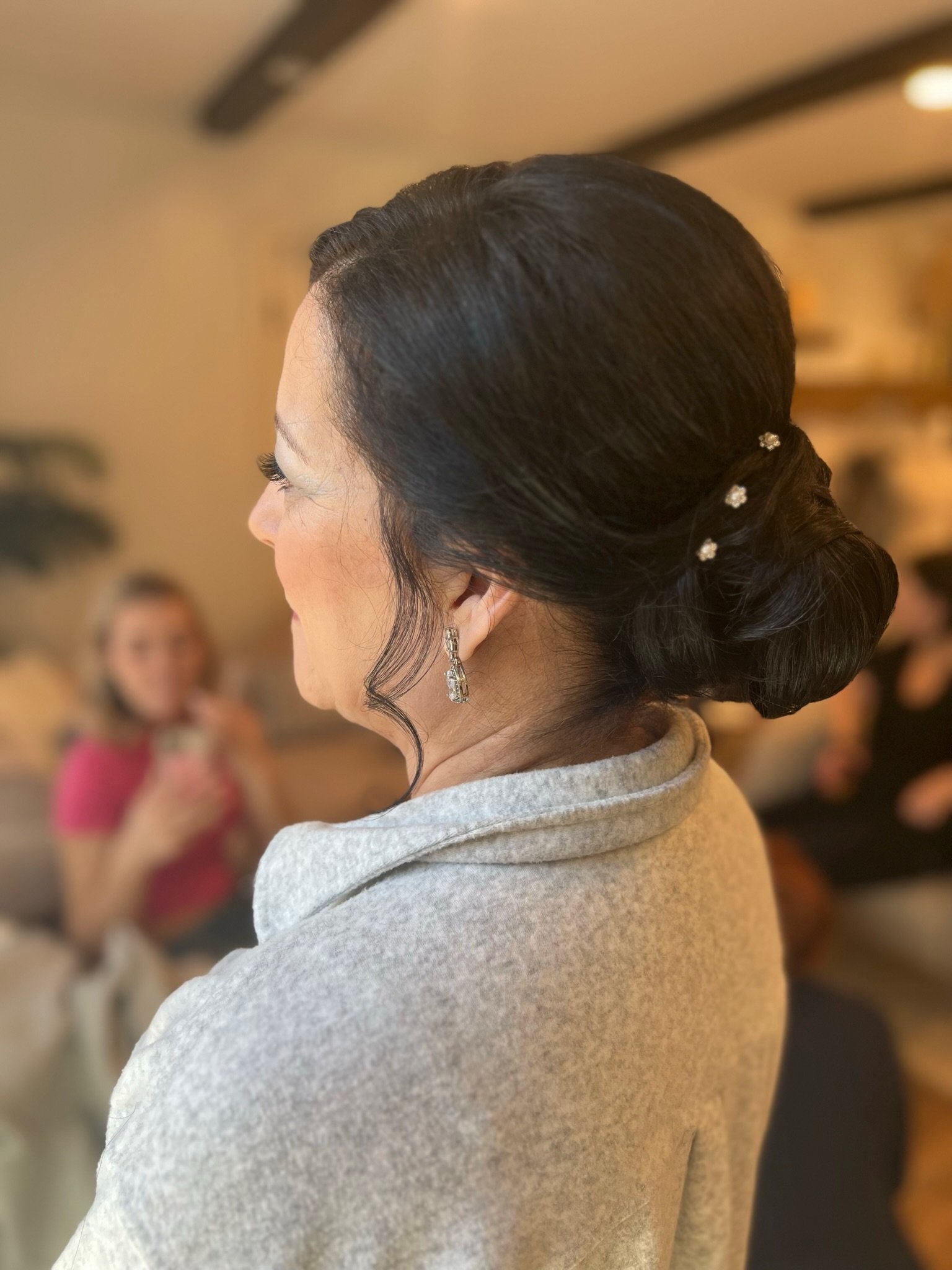 A woman with dark, styled hair decorated with small pearl or crystal hairpins, wearing earrings and a gray shawl, at an indoor event with blurred people in the background.