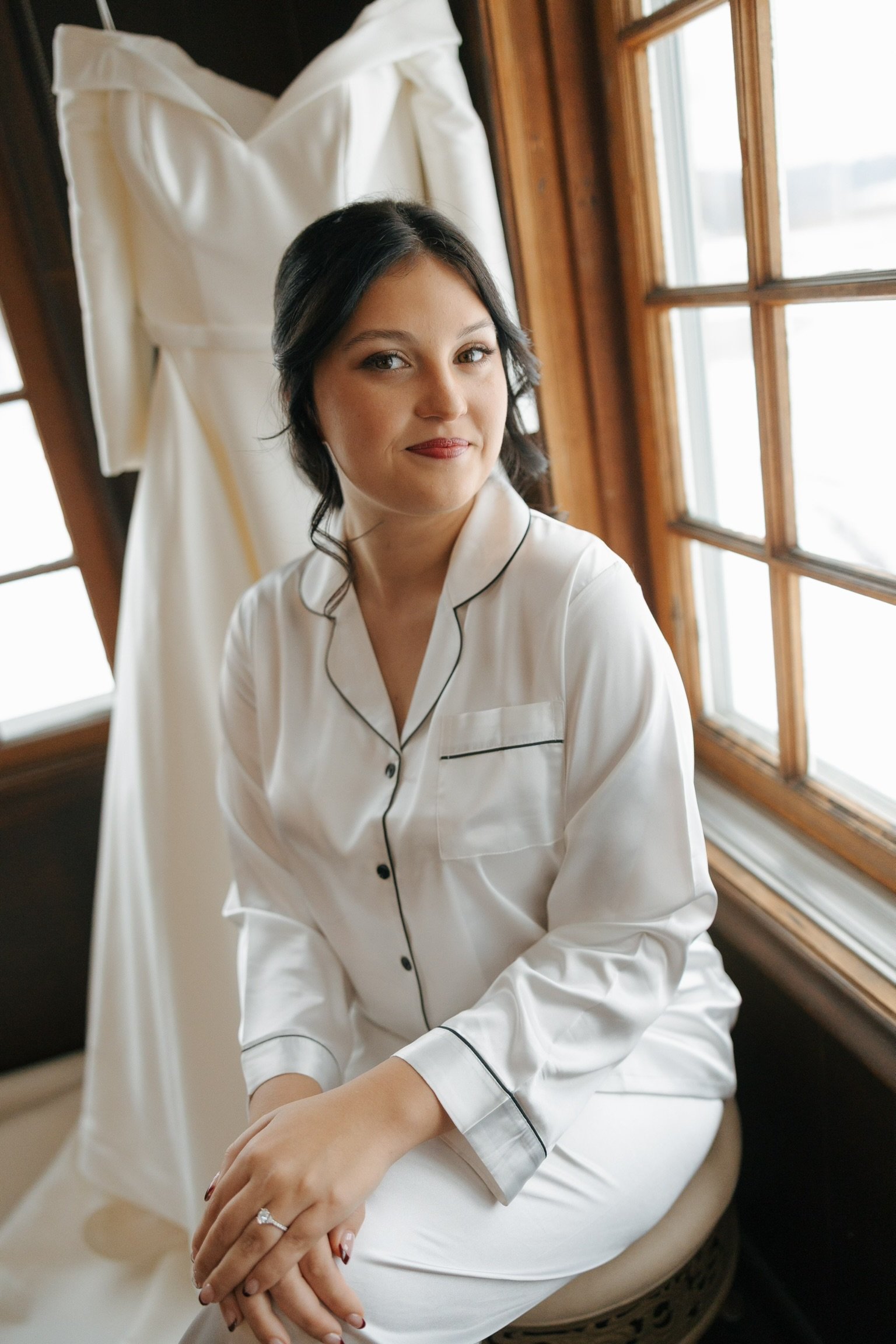 A young woman with dark hair and light skin wearing white satin pajamas with black piping, sitting near a window with wooden framing, a wedding dress hanging behind her, her hands resting on her lap, with a ring on her left ring finger.