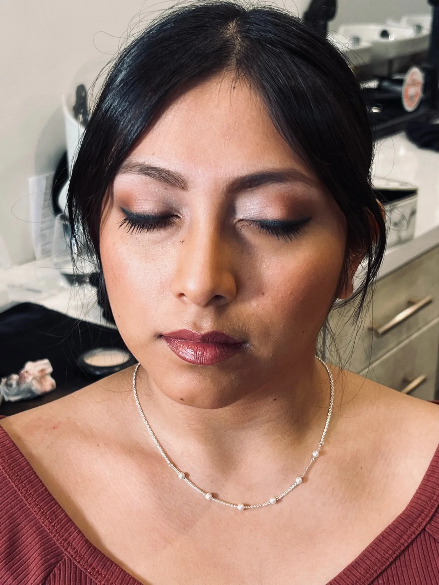 A woman with closed eyes and dark hair styled with a side part, wearing makeup including eyeliner and lipstick, a silver necklace with pearls, and a red off-the-shoulder top, standing in what appears to be a salon or makeup studio.