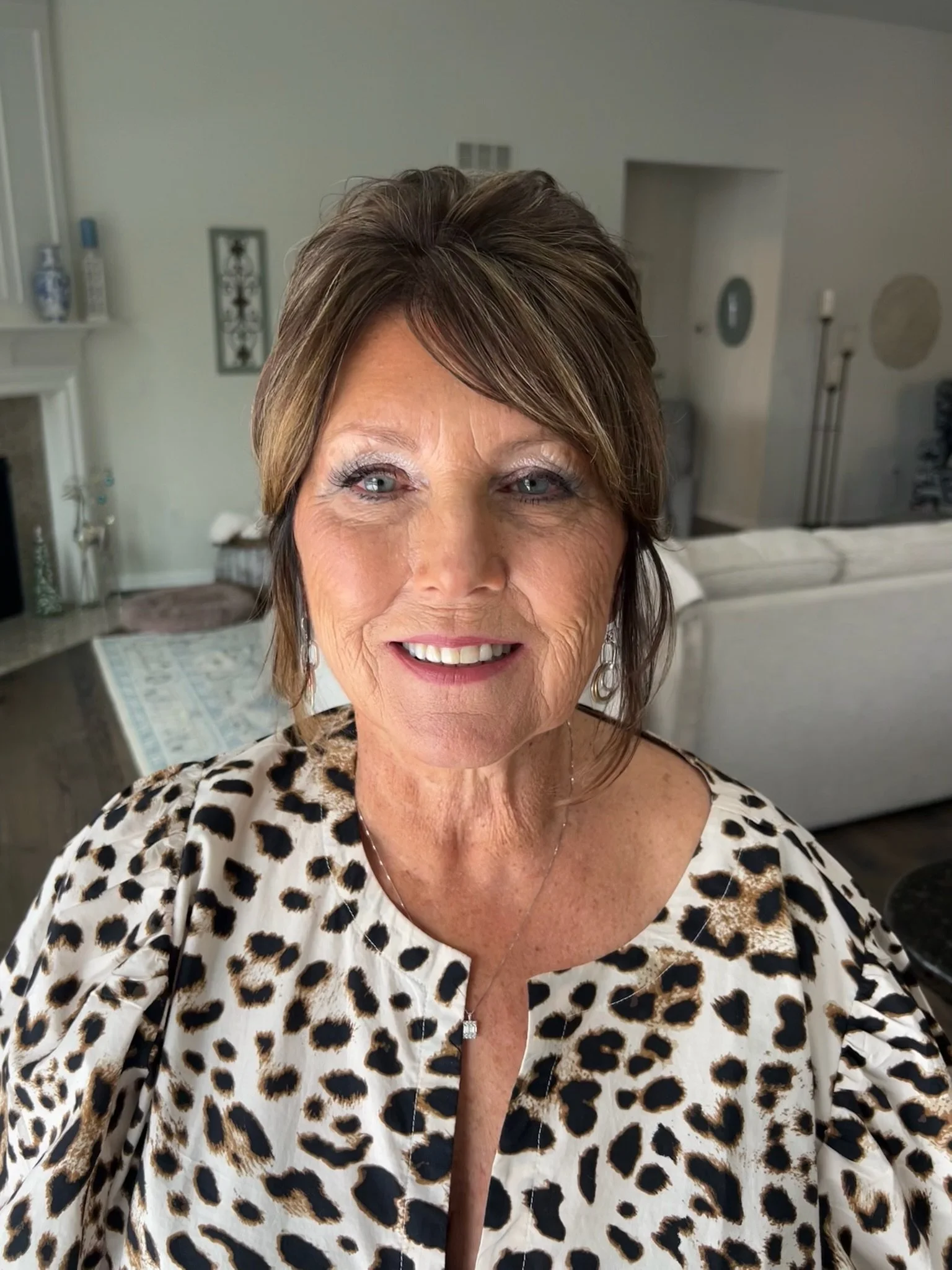 Close-up of a smiling woman with short brown hair and wearing a leopard print top, standing in a living room with light-colored walls, a white couch, and decorative items in the background.