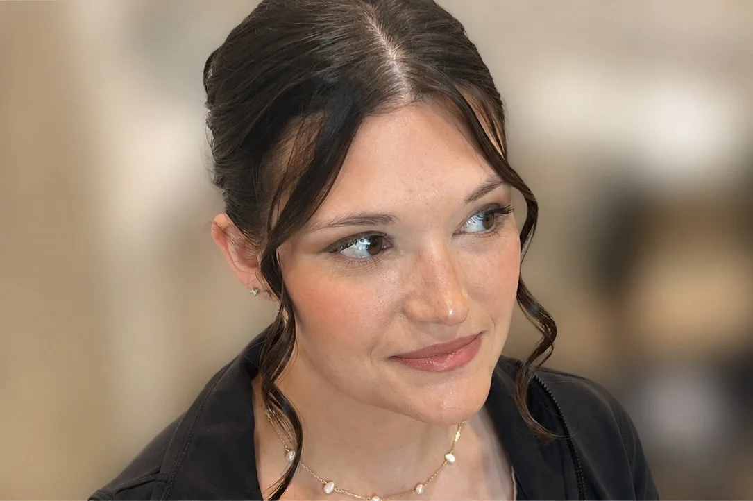 Close-up of a young woman with dark brown hair styled in loose curls, wearing makeup, small earrings, a black jacket, and a delicate necklace, looking slightly to the side with a soft smile.