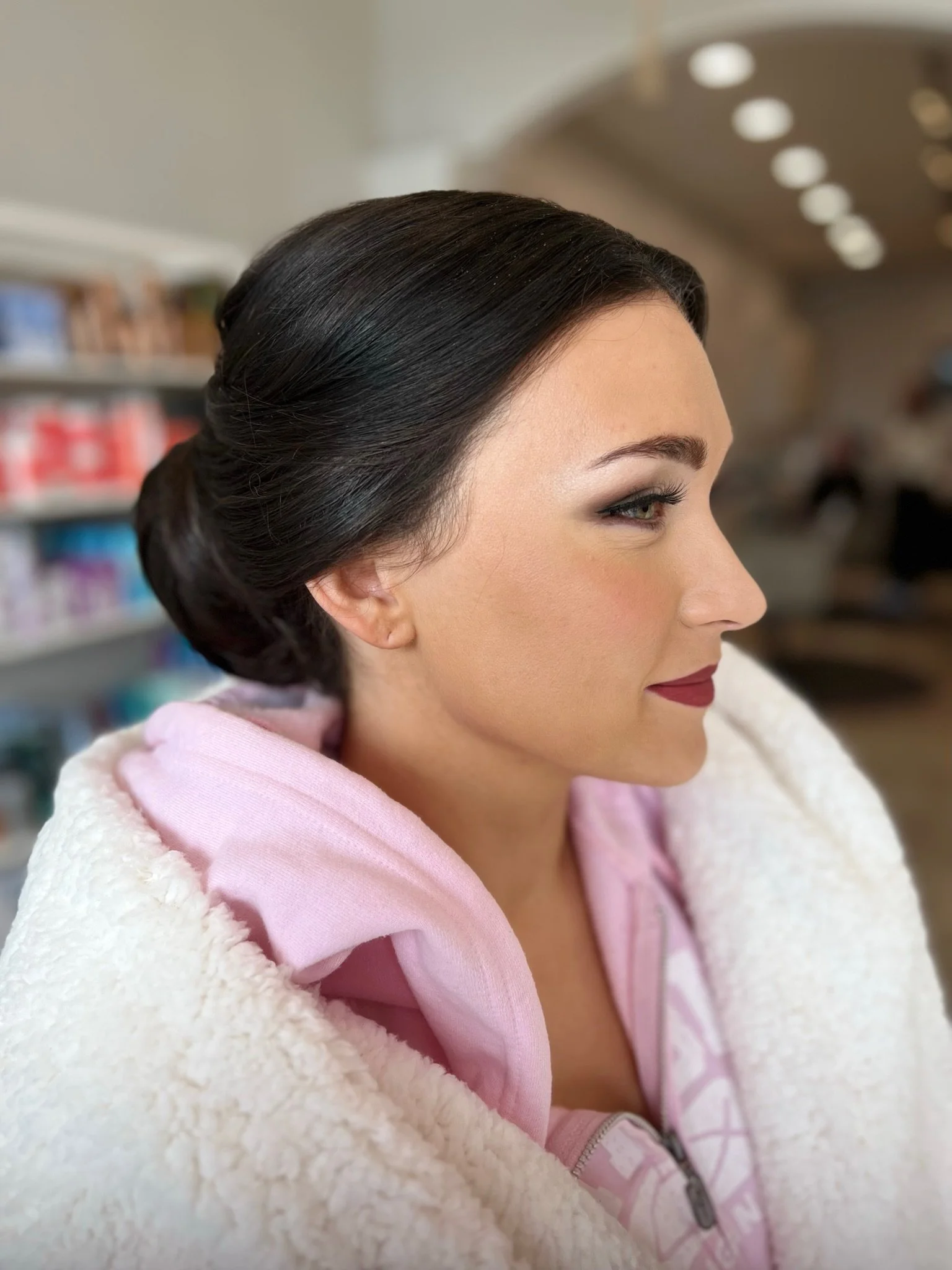 Side profile of a woman with dark hair styled in an elegant updo, wearing a pink hoodie and a white fluffy jacket, in a store with shelves in the background.