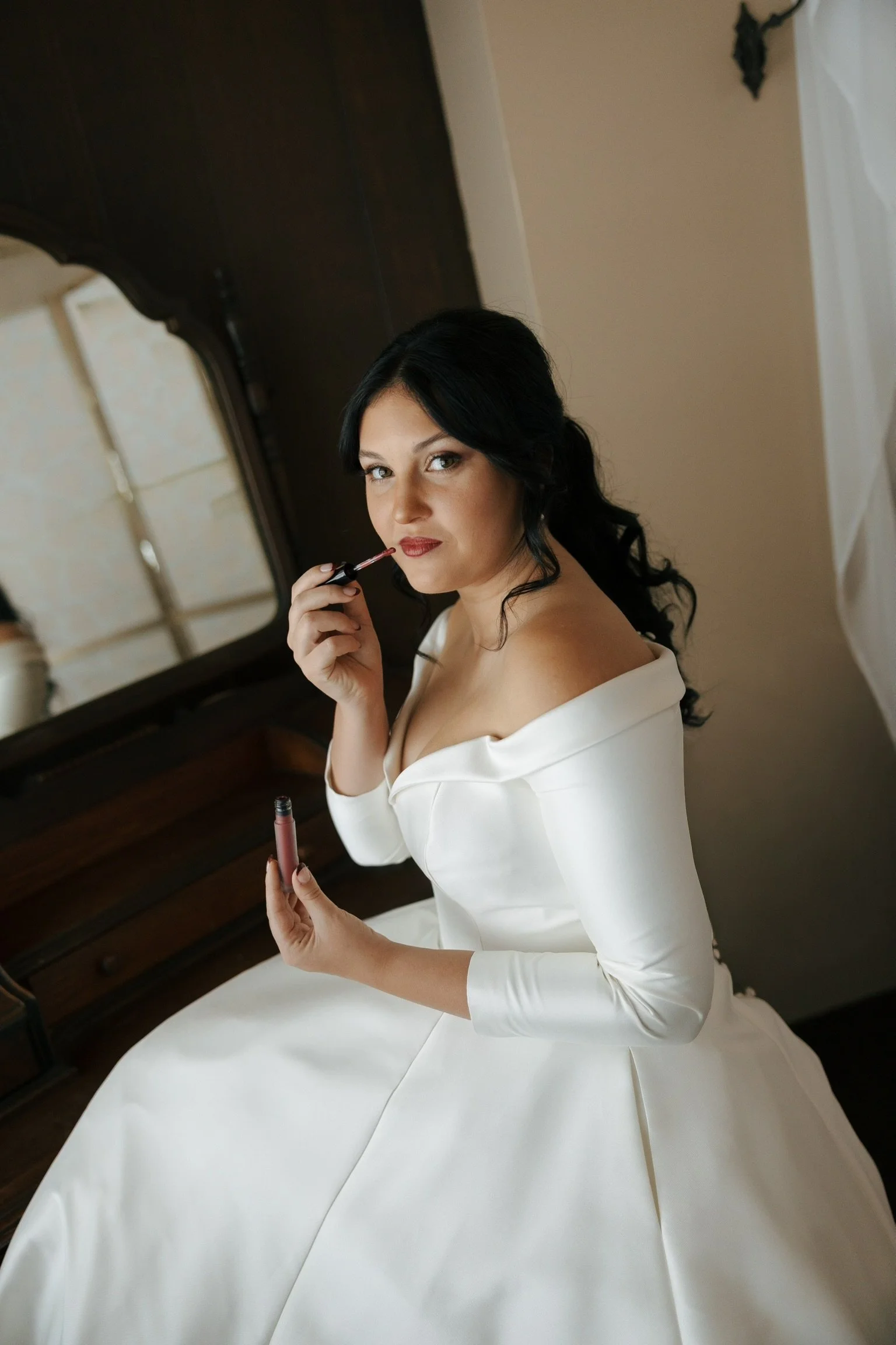 A woman with black hair and blue eyes wearing a white off-shoulder dress, sitting on a bed and applying red lipstick in front of a mirror.