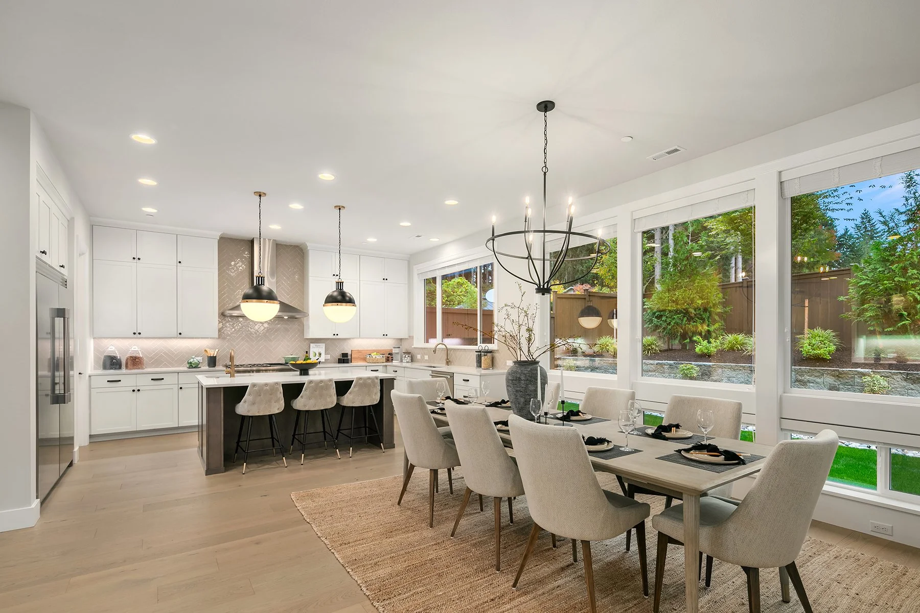 Modern open-concept kitchen and dining area with white cabinetry, a kitchen island with barstools, large windows showing a garden, and a dining table set with black placemats, glasses, and cutlery.