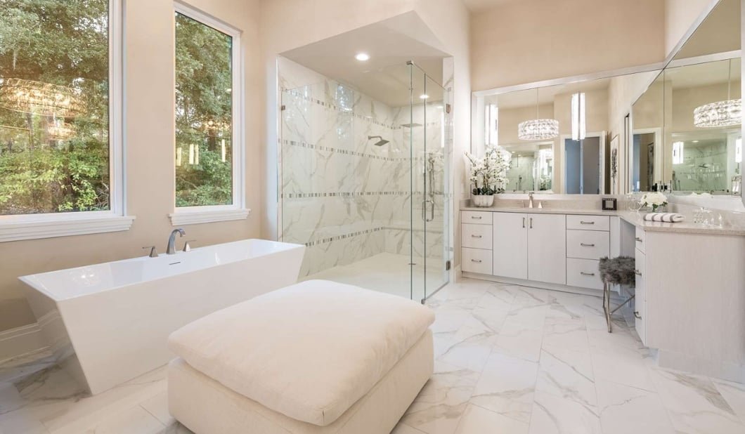 Bright, modern bathroom with a white freestanding bathtub, large windows, marble floors, a walk-in shower with glass doors, white cabinets, and a large mirror.