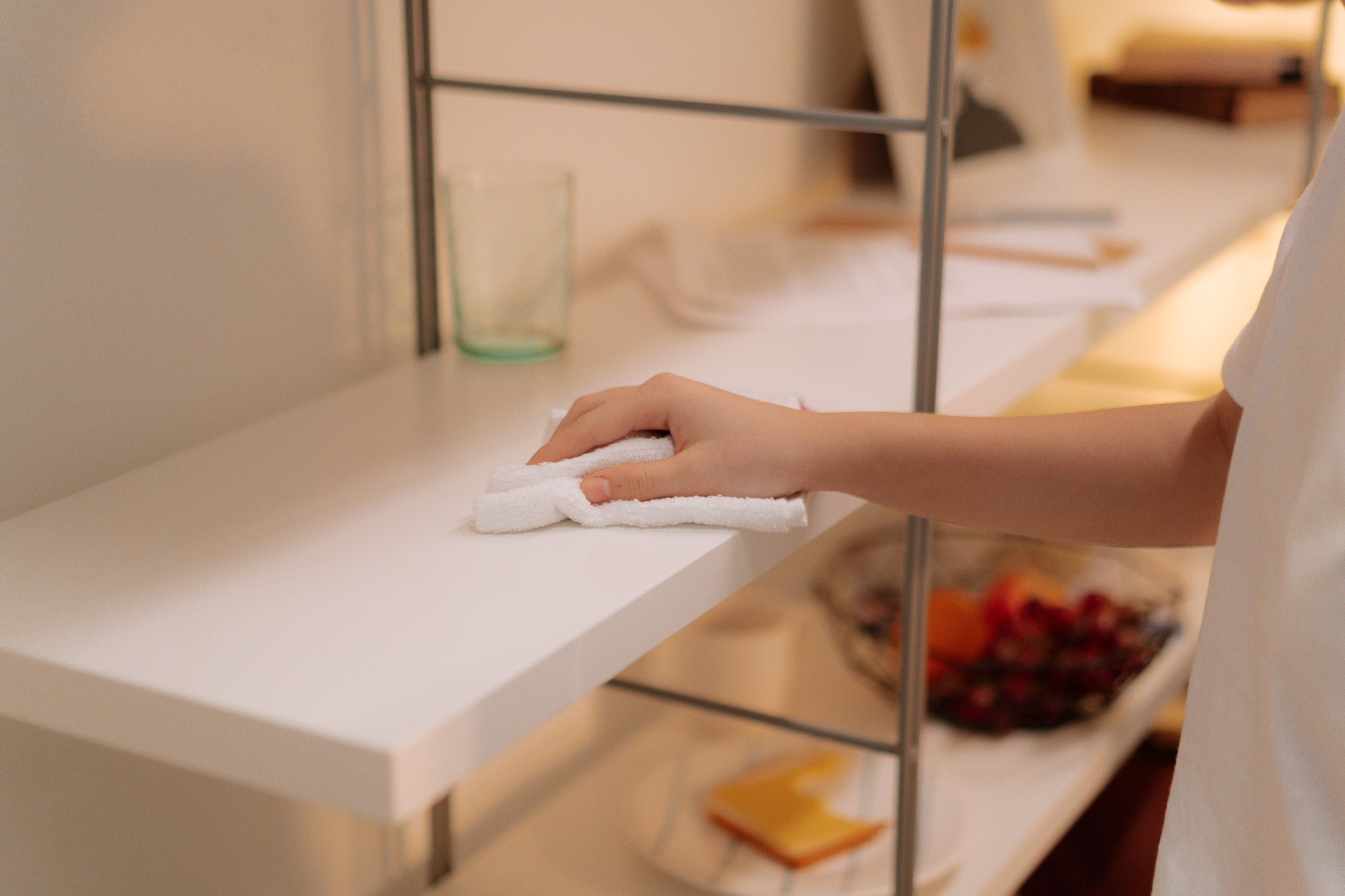 Person wiping a white shelf with a cloth, with various glassware and books on the shelves.