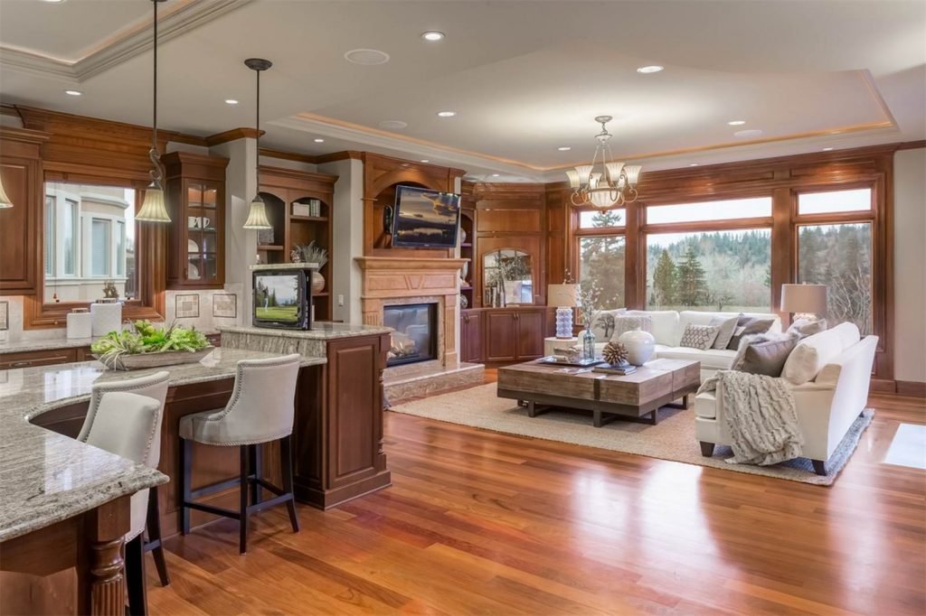 A spacious living room with large windows, hardwood floors, white sofa, wooden coffee table, fireplace, and TV, with a kitchen counter and pendant lights visible on the left.