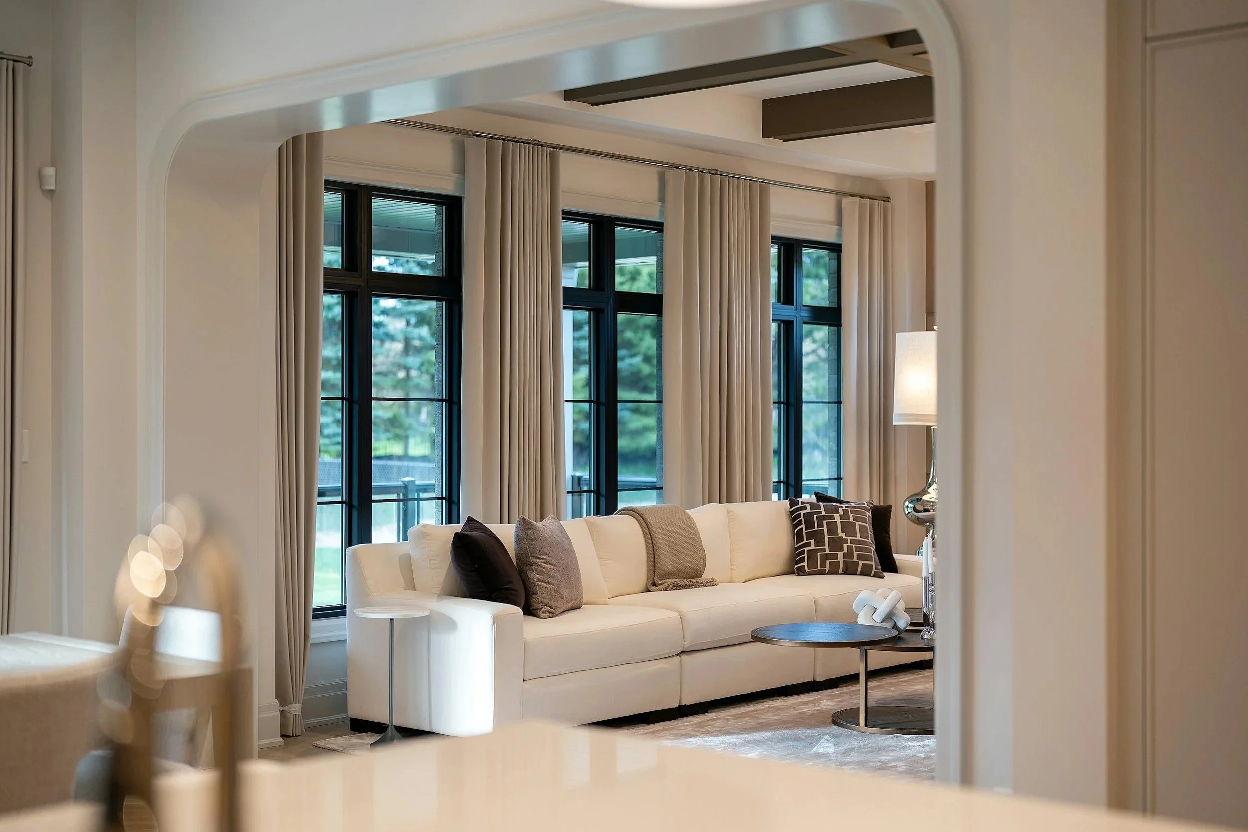 Living room with beige walls, large black-framed windows with white curtains, a white sectional sofa with multiple decorative pillows, a small side table, and a modern table lamp.