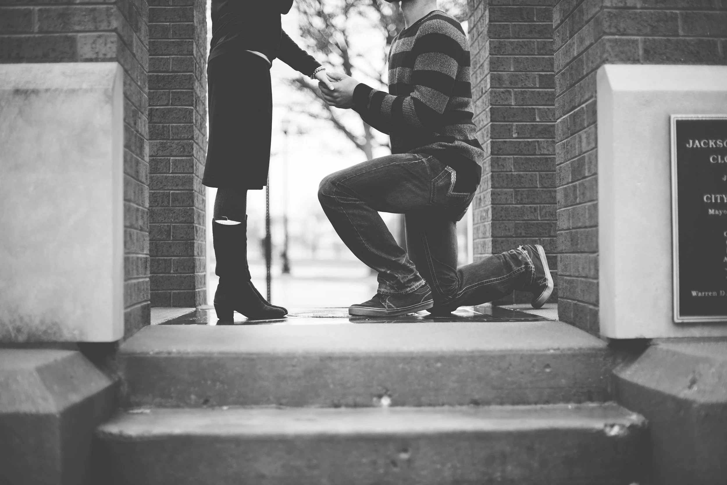 A man kneeling on one knee, proposing to a woman outside between brick walls, holding her hand. The woman is standing, wearing high heels, dark clothing, and ripped jeans. The scene is captured in black and white.