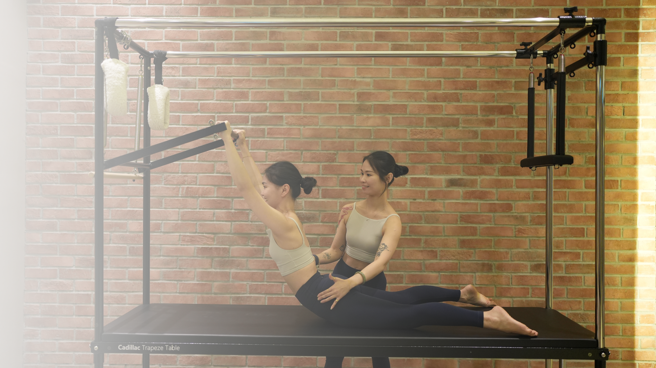 Two women practicing yoga on a trapeze table against a brick wall. One woman is holding a yoga pose while the other assists her.
