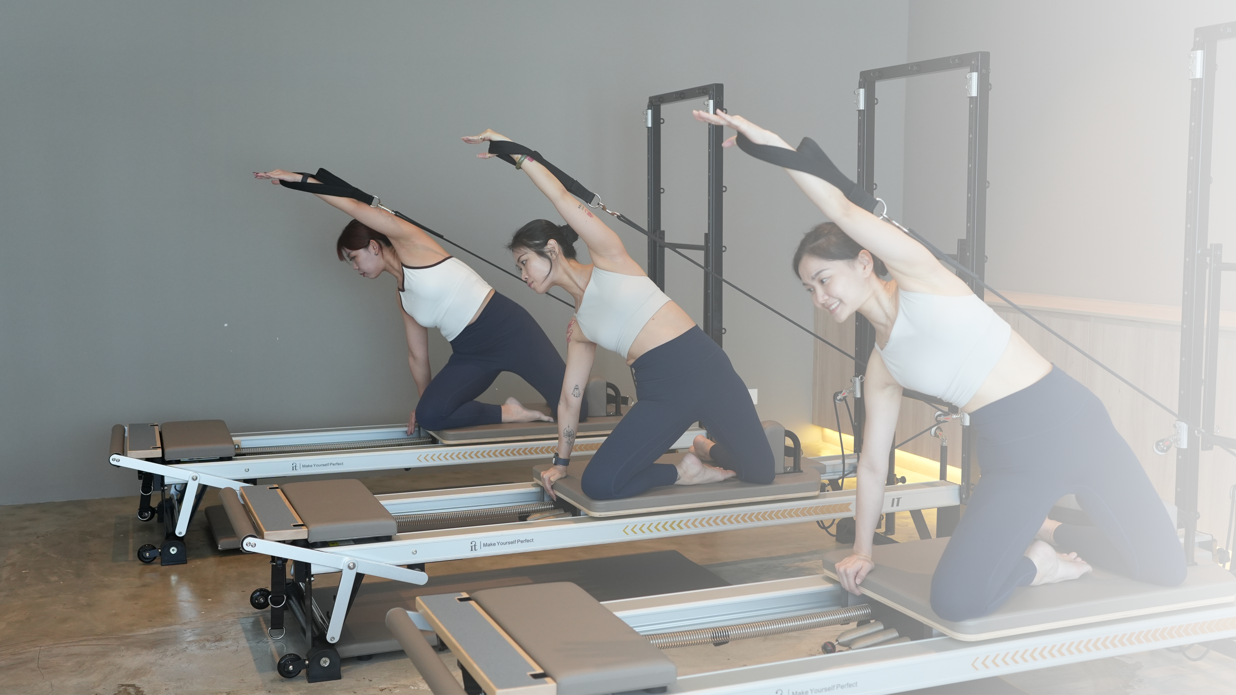 Three women in workout attire on reformer Pilates machines performing side stretches with arms extended overhead, in a fitness studio with gray walls.