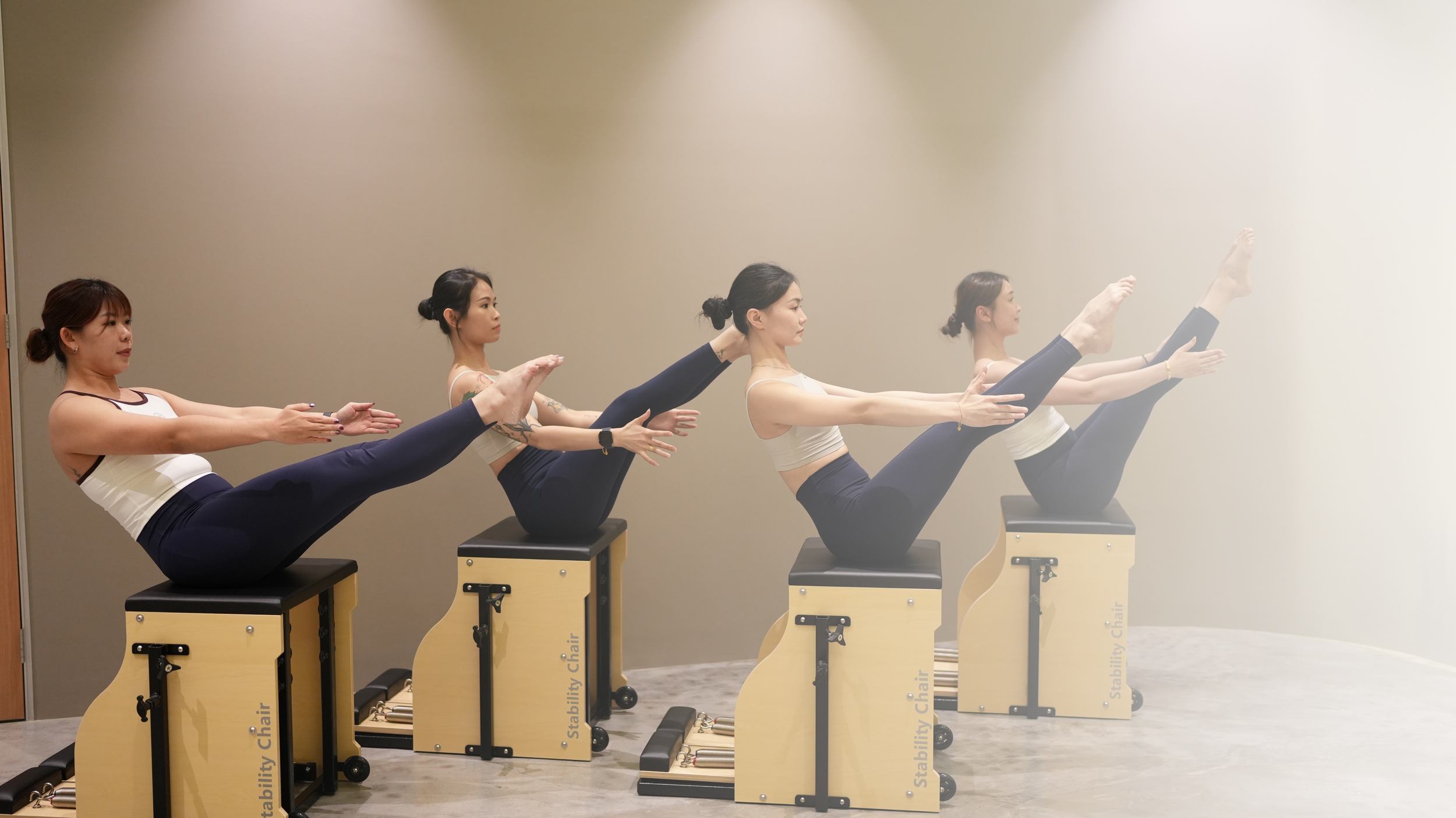 Five women practicing yoga in a seated balancing pose on wooden exercise chairs, with legs raised and extended forward, arms extended straight, and focused expressions.