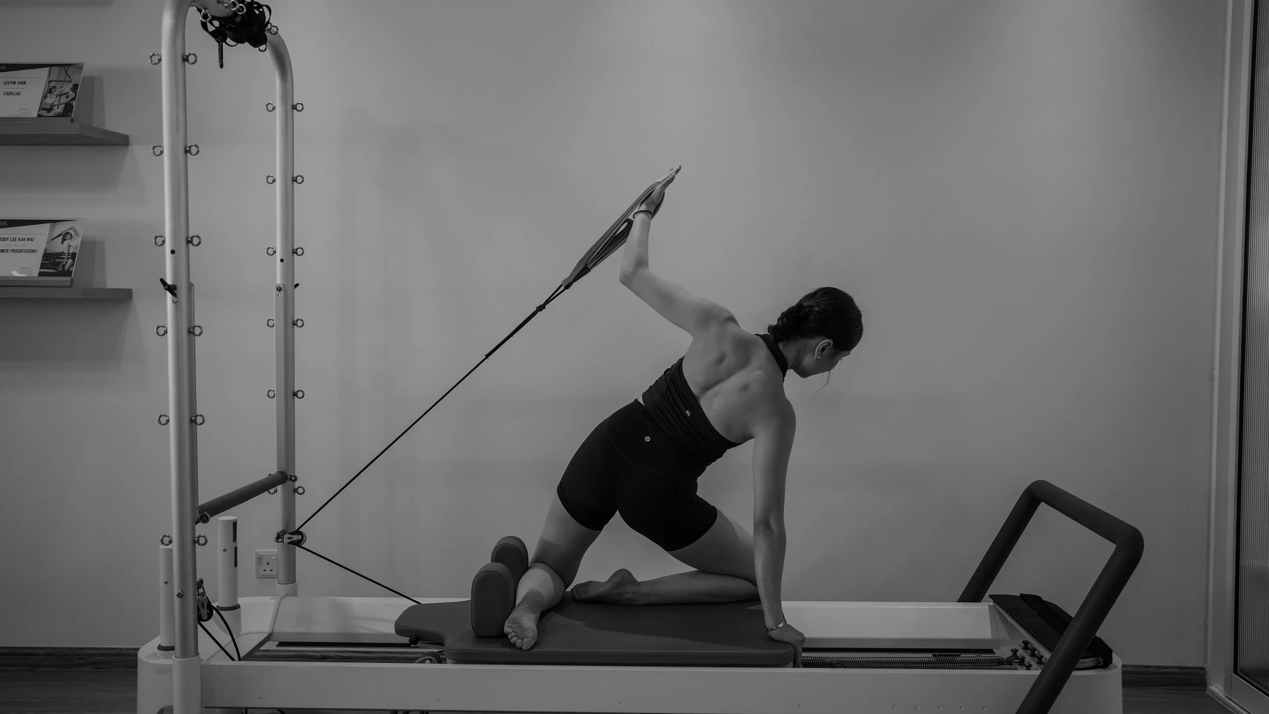 A woman exercising on a Pilates reformer machine, leaning to one side with one arm extended overhead and the other hand on the mat, in a fitness studio.