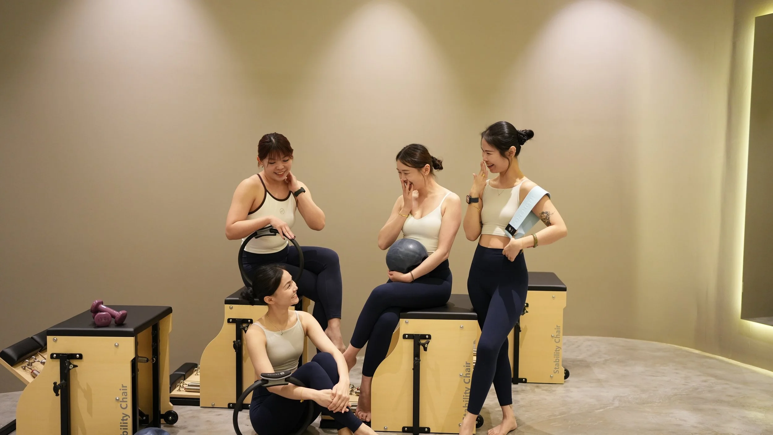 Four women in workout attire at a fitness class engaging in a fitness activity with stability chairs and a medicine ball in a gym A0.