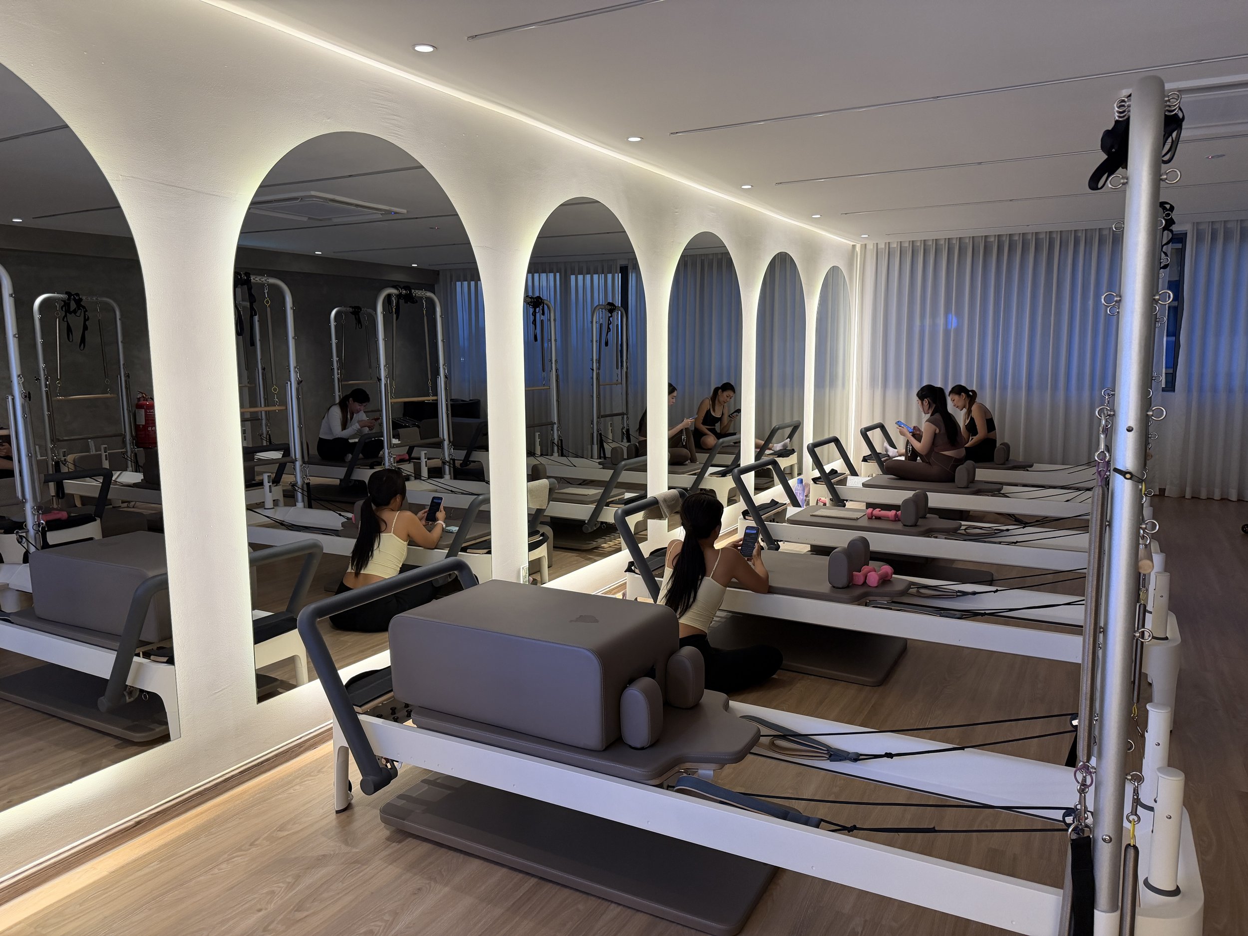 A modern fitness studio with Pilates reformer machines arranged in rows. The studio has white arched pillars, wood flooring, and a ceiling with recessed lighting. Several women are sitting or using their phones, and there is a curtain with vertical stripes in the background.
