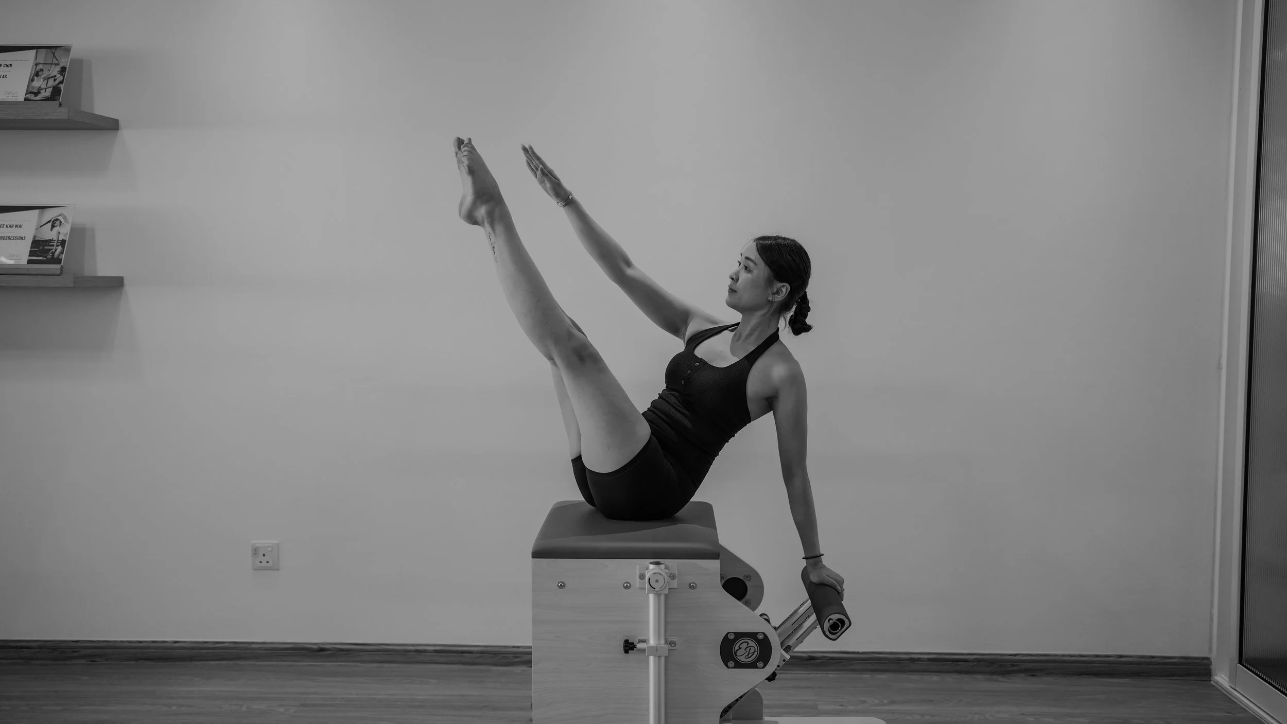 A woman performing a Pilates exercise on a reformer machine in a gym, balancing with one leg raised and arms extended.