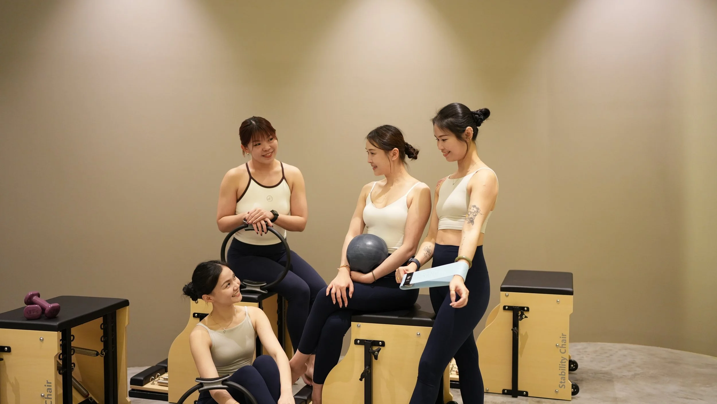 Five women in workout clothes are talking and smiling in a gym or fitness class setting, with some sitting and others standing, surrounded by workout equipment like dumbbells and a stability chair.