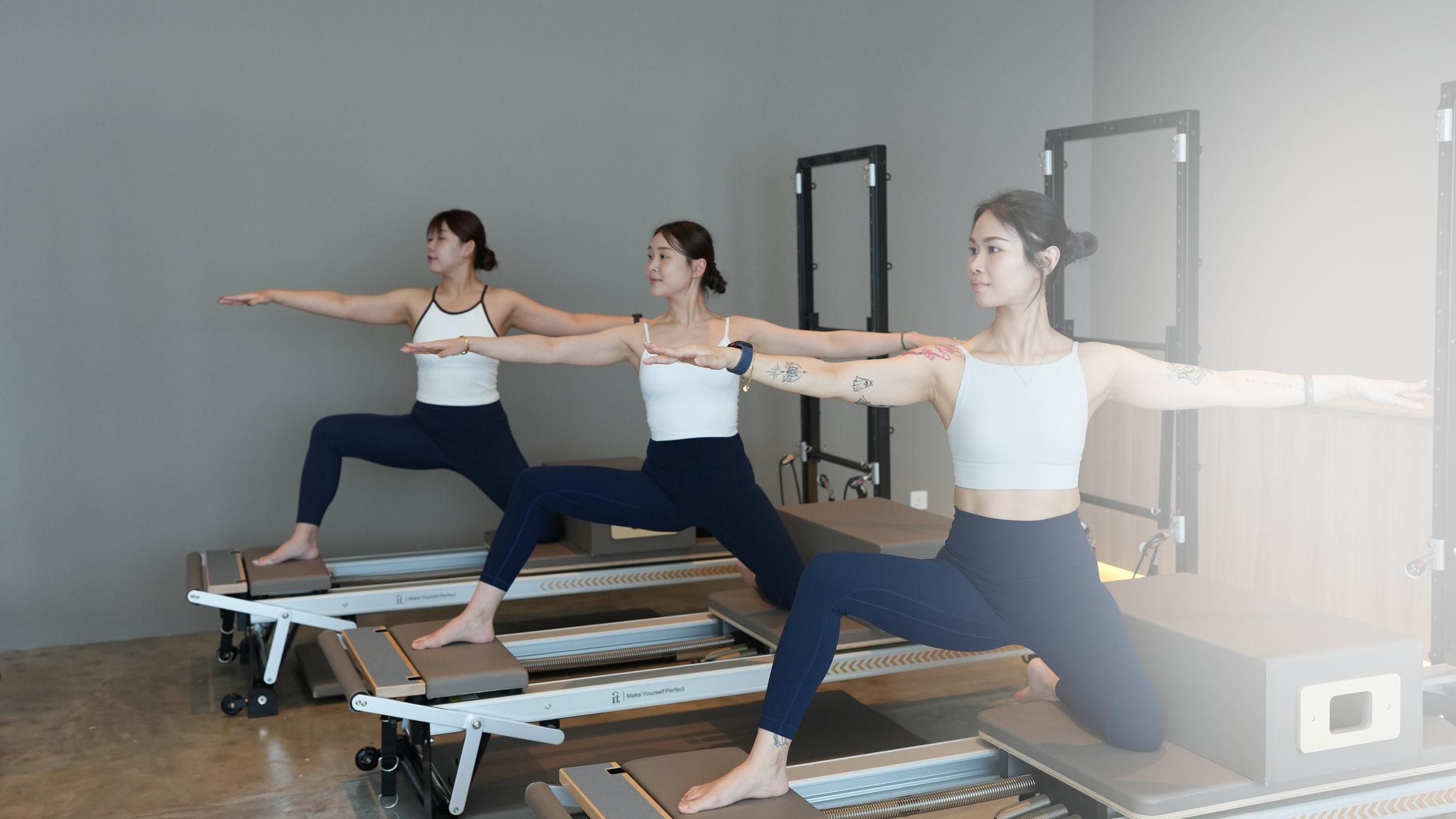 Three women practicing Pilates in a studio, positioned on reformer machines, extending arms forward and balancing on one leg.