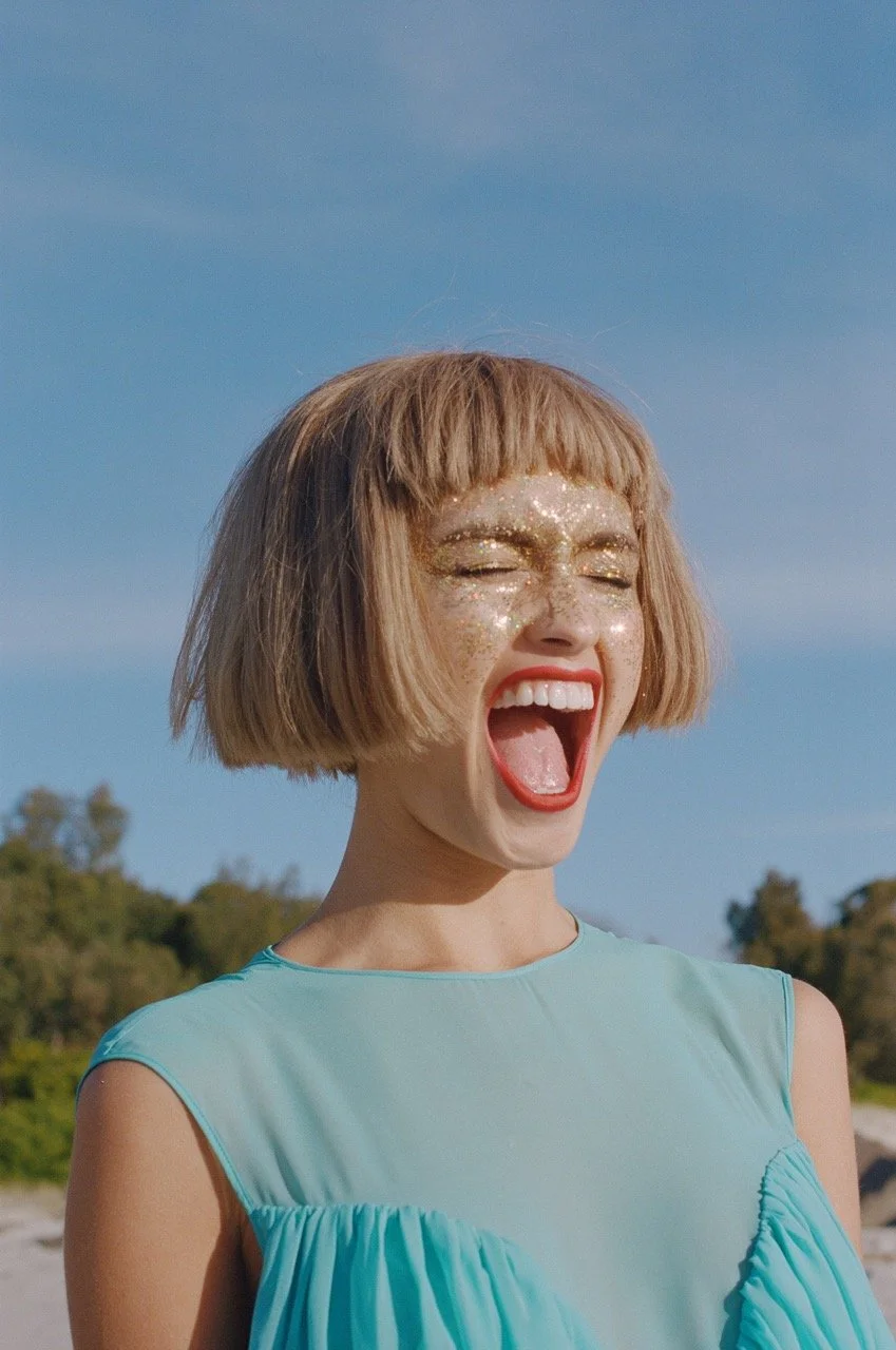 Woman with short blond hair, glitter on her face, open mouth, tongue out, wearing a sleeveless light blue dress outdoors under blue sky.