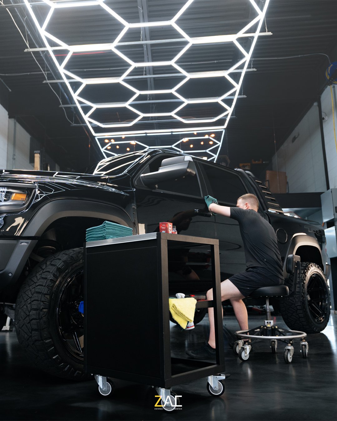 A person cleaning a black pickup truck inside a modern workshop with hexagonal ceiling lights.