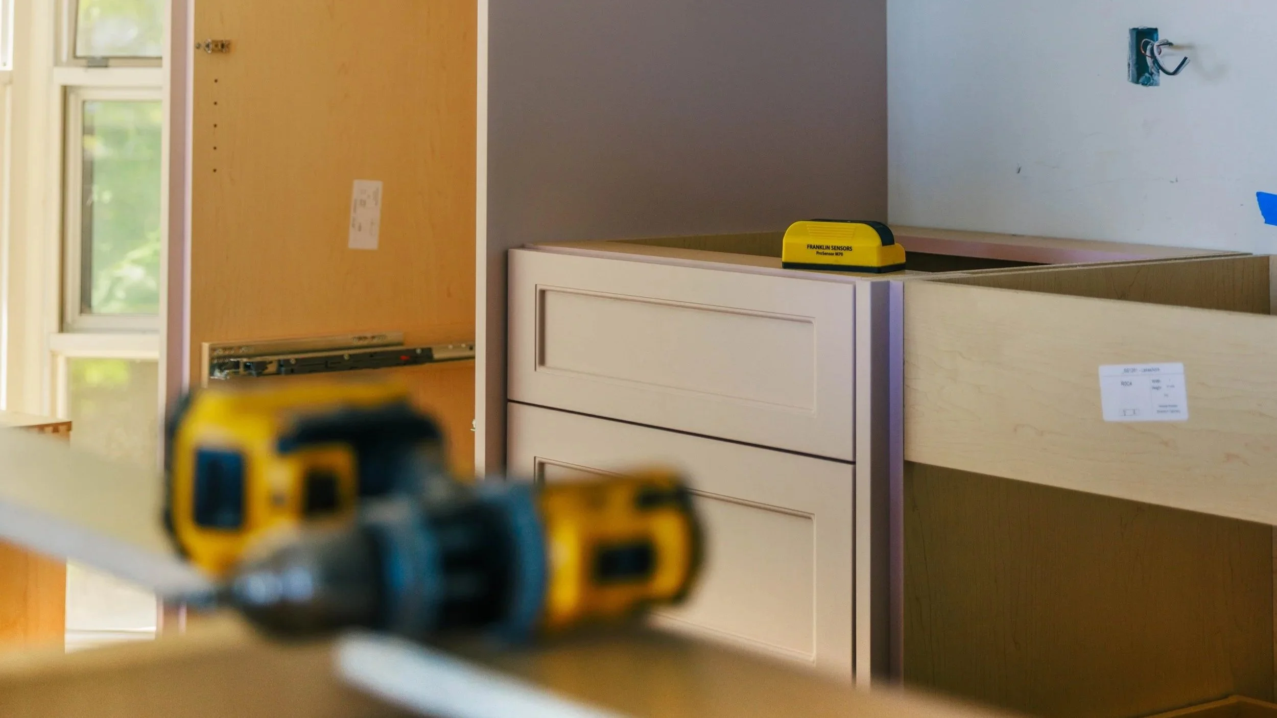 Construction tools in the foreground with a partially assembled white dresser in the background, inside a room with windows and light-colored walls.