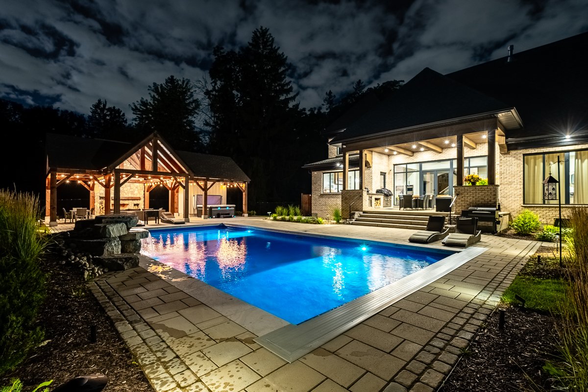Night view of a lit backyard with a swimming pool, house, and outdoor seating area.