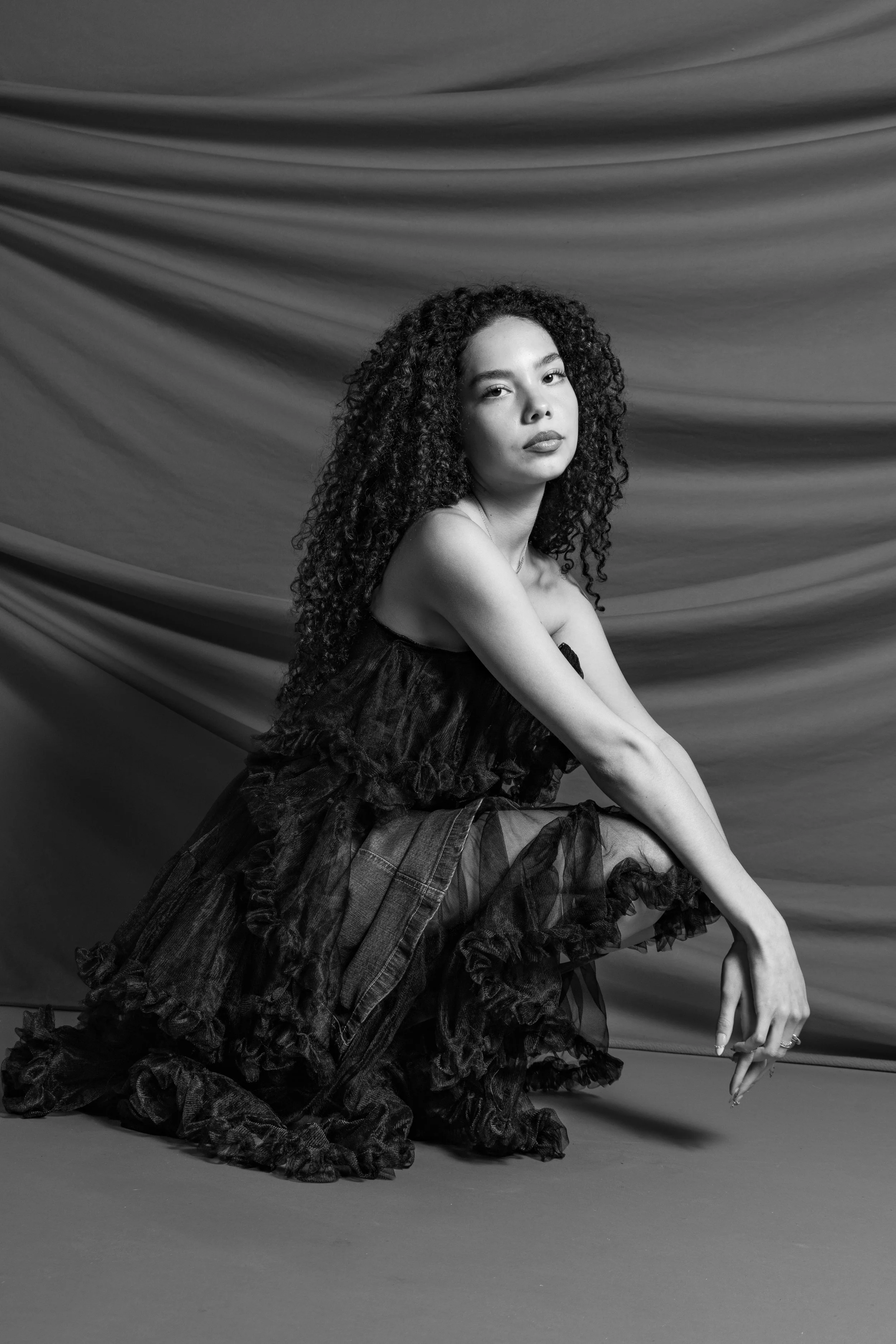 A woman with curly hair sitting on the floor in front of a textured backdrop, wearing a layered, ruffled dark dress, with her arms crossed over her knees.
