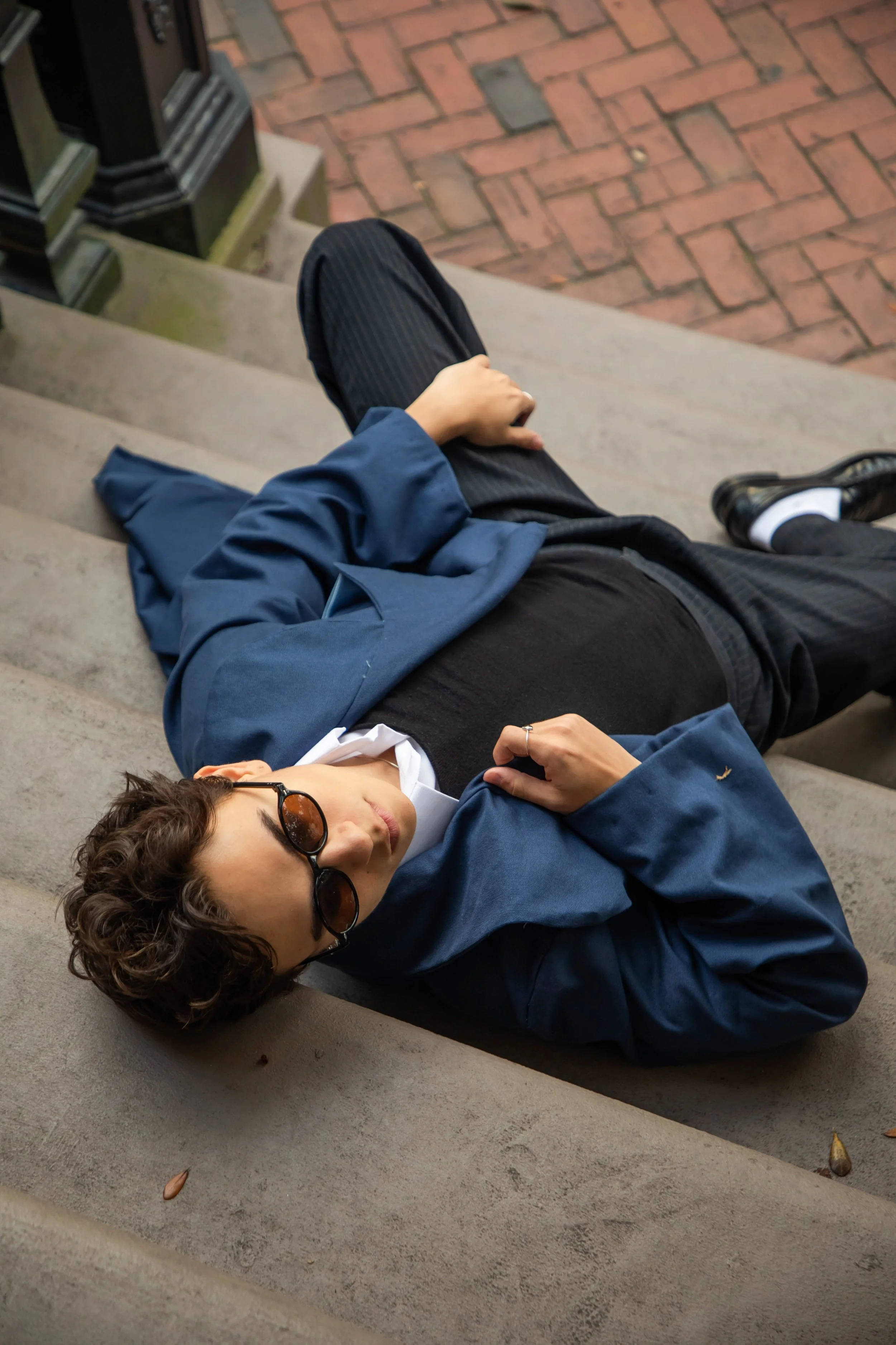 A young man with curly hair and sunglasses lying on a concrete bench, wearing a blue jacket, black pants, and black shoes. There are steps and a brick sidewalk nearby.