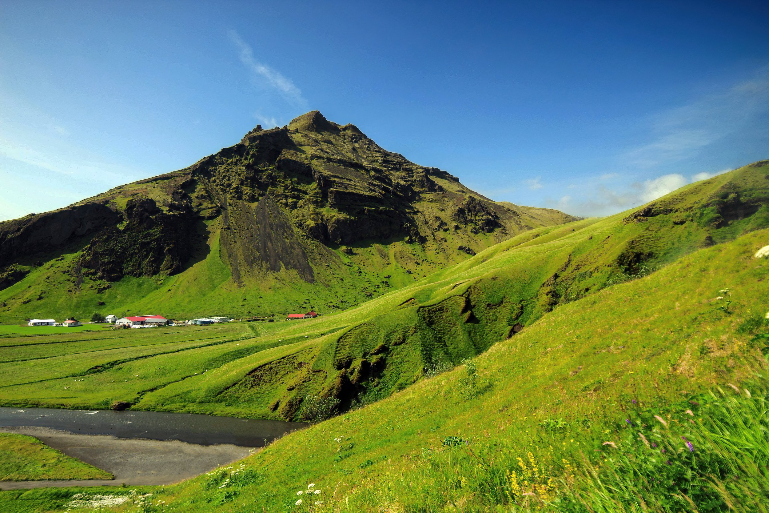 Green grassy hillside with a mountain and small houses at the base, under a blue sky with a few clouds.