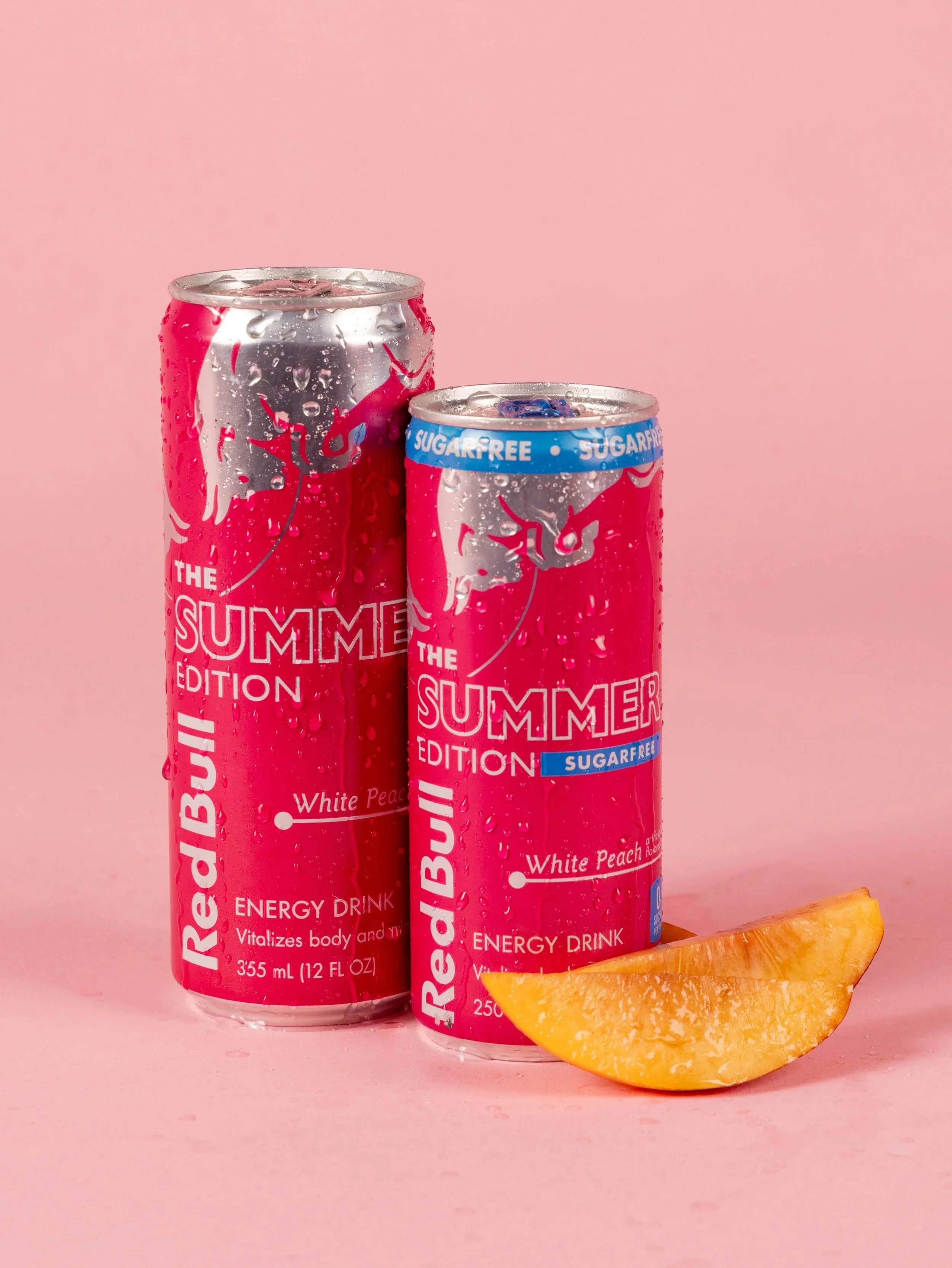 Two cans of Red Bull Sugarfree energy drink with a slice of orange on a pink background, water droplets on the cans.