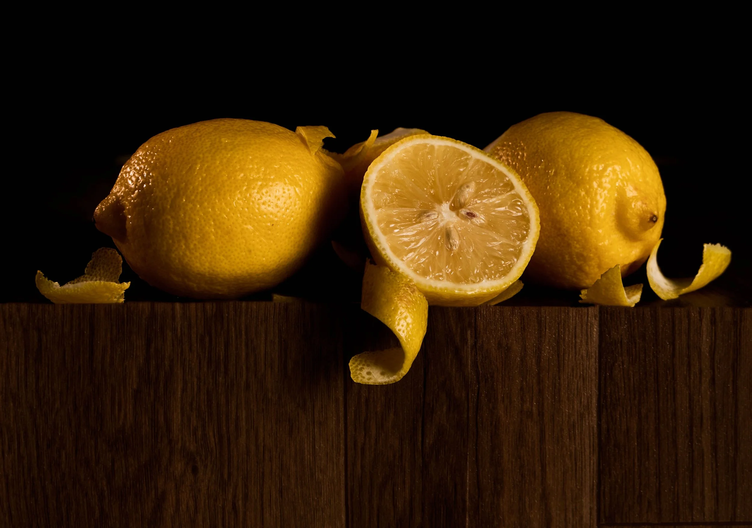 Three lemons, one sliced in half, resting on a wooden surface with lemon peels scattered around, against a dark background.