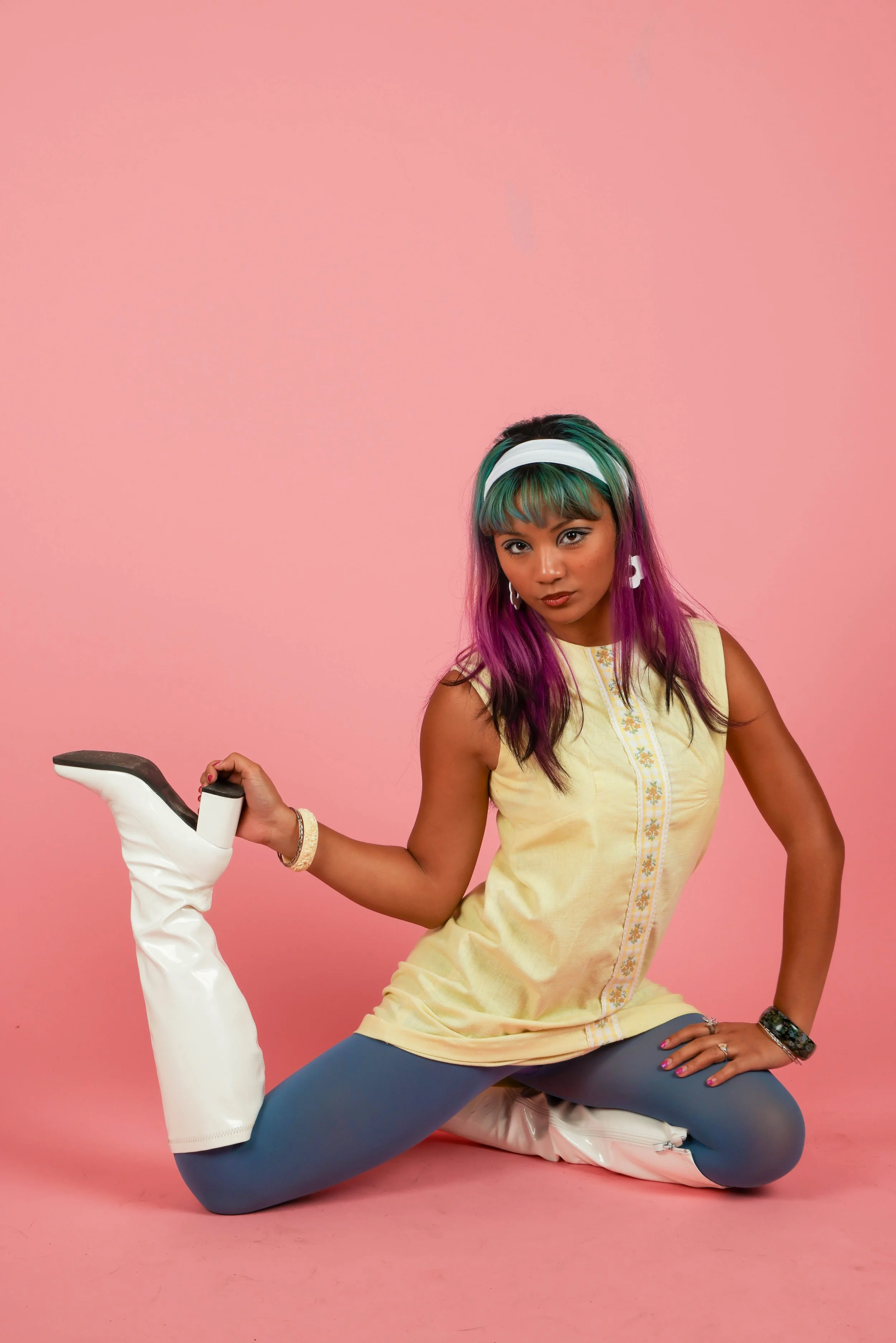 A woman with colorful hair, wearing a yellow embroidered top, gray leggings, and white over-the-knee boots, posing against a pink background.