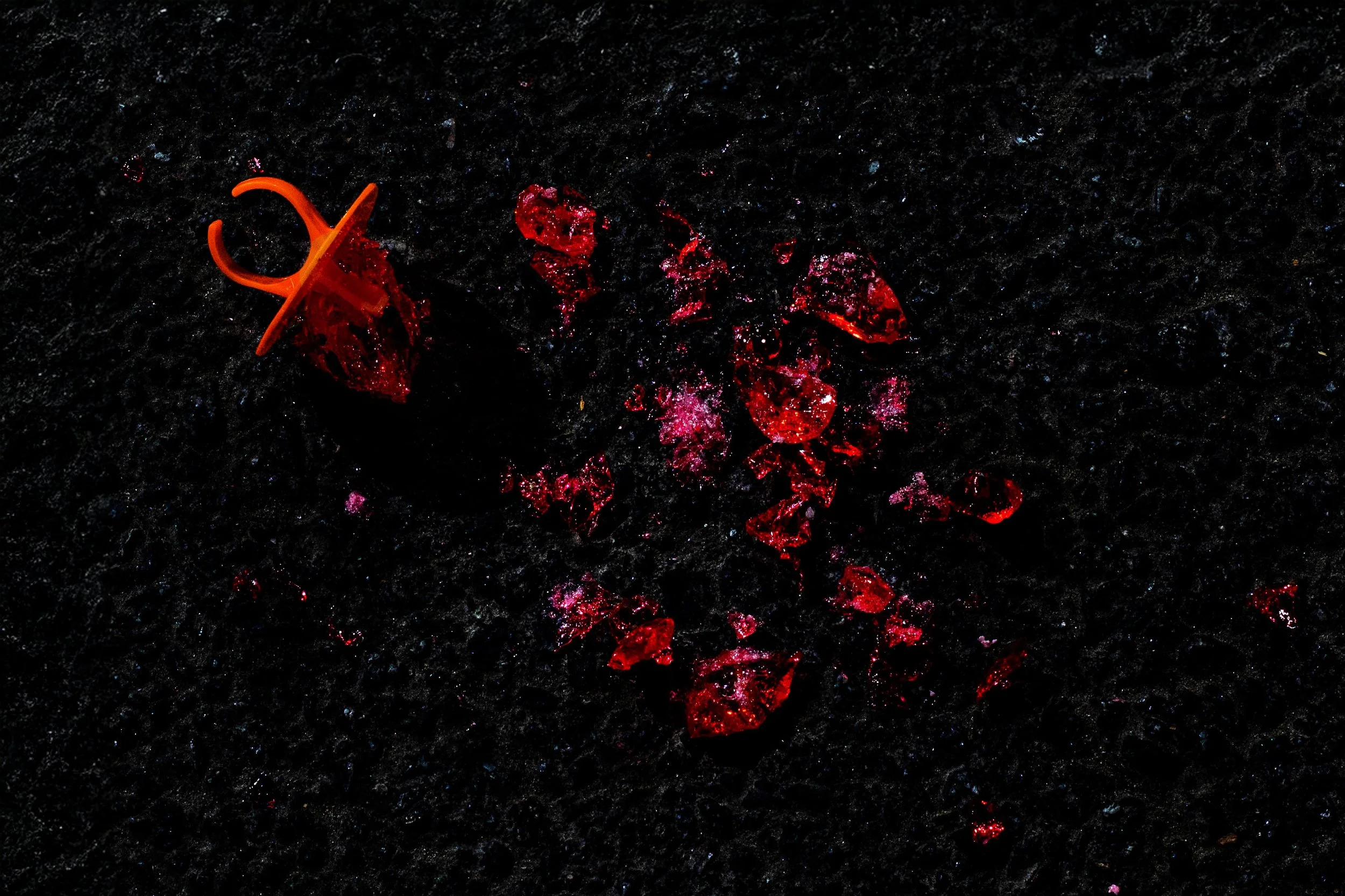 Broken red glass pieces and an orange plastic hook on a dark, textured surface with reflections.