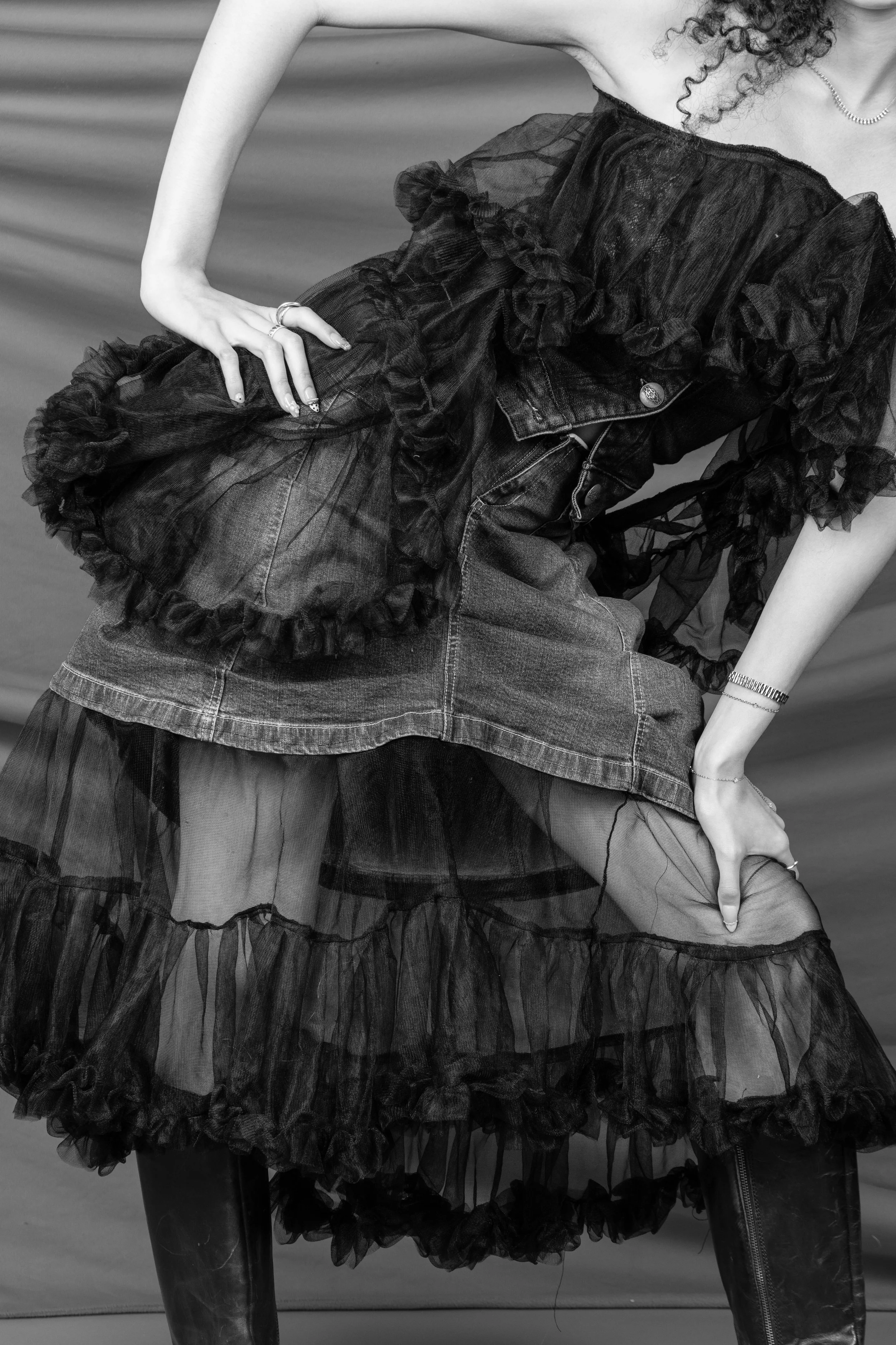 Close-up of a woman in high fashion clothing, wearing a layered tulle skirt, denim shorts, rings, a pearl necklace, and a watch, with curly hair, against a plain background.