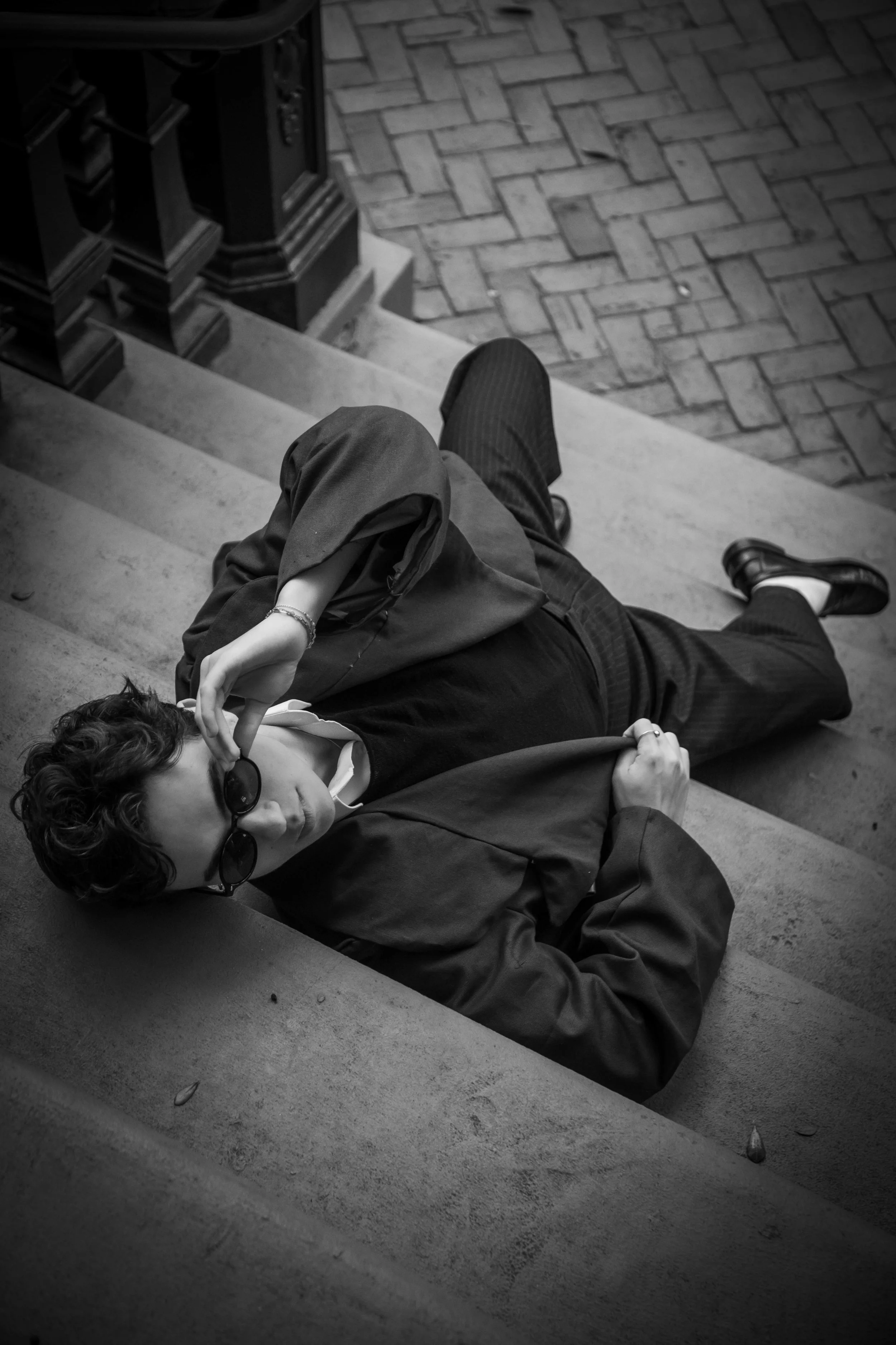 A person reclines on concrete stairs with a brick pavement below, wearing sunglasses, a dark jacket, and patterned pants, with one arm behind their head and the other resting on their chest.