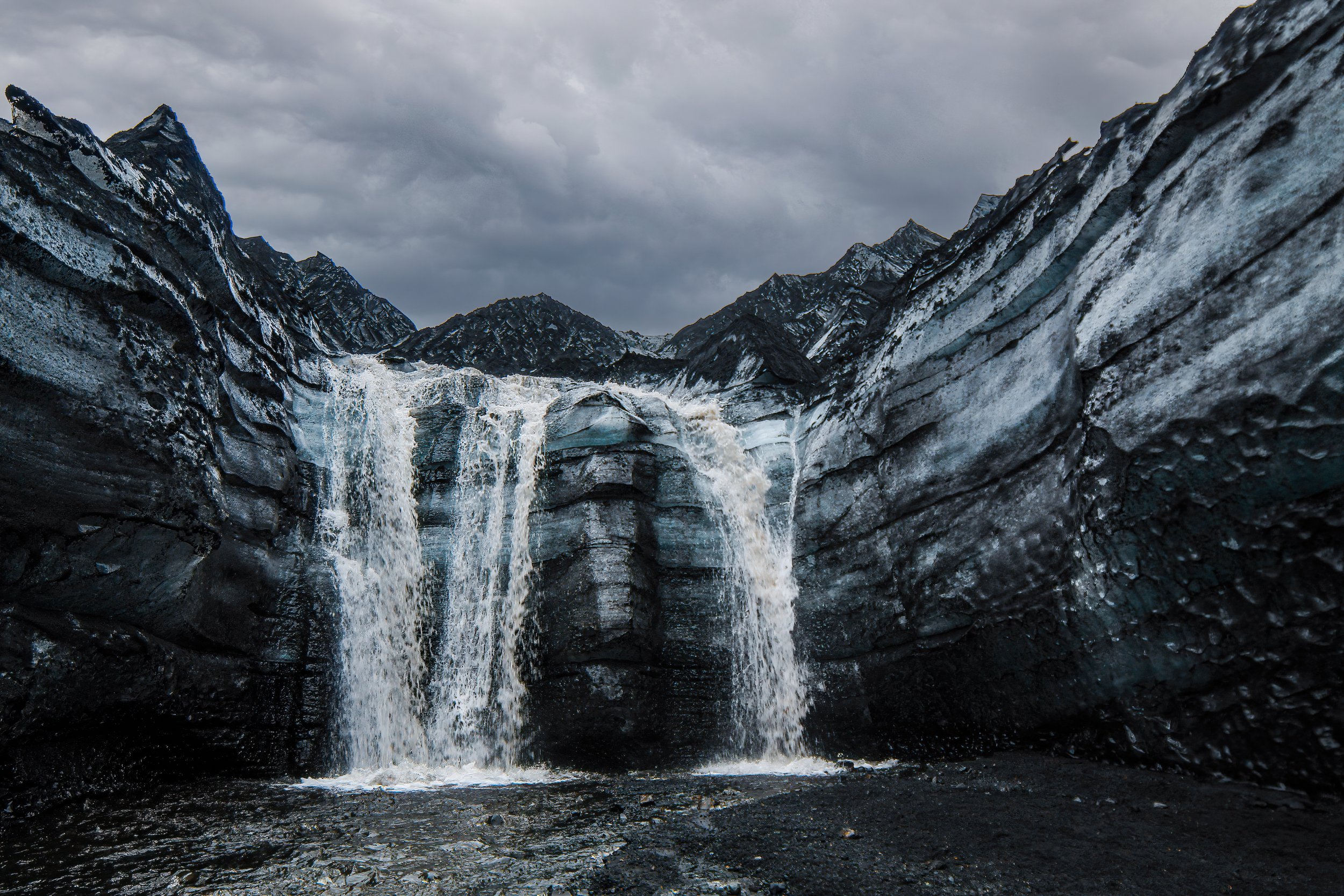 A rugged mountain landscape with dark, jagged rocks and a waterfall flowing into a pool, under a cloudy sky.